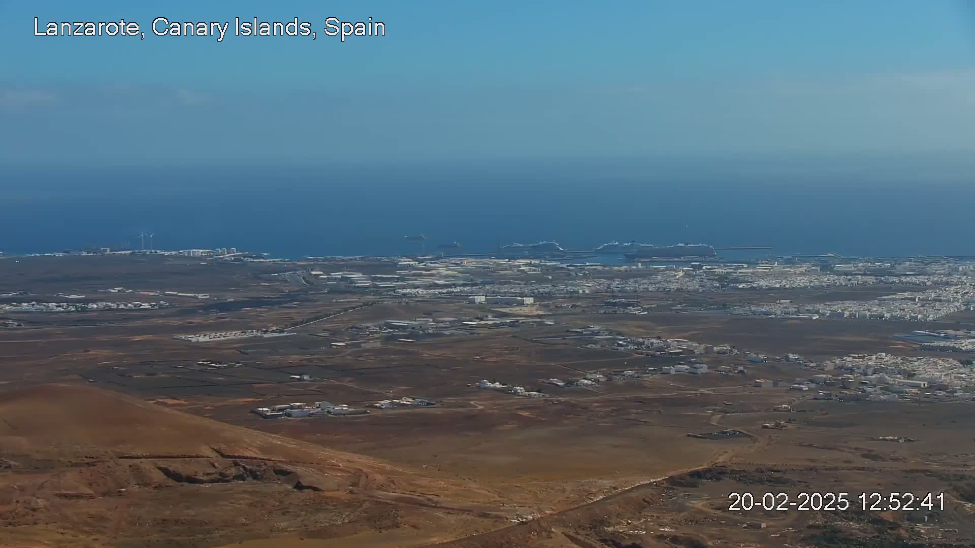 A high-altitude view shows a dry, brown landscape dotted with low buildings extending to a coastline with a port and calm blue ocean under a clear sky.