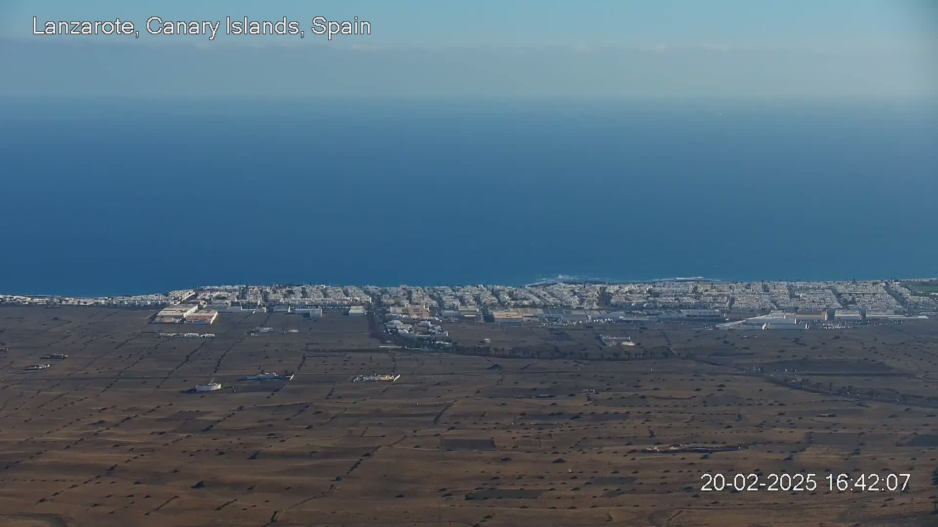 An aerial view shows a coastal town with light-colored buildings next to a dark brown, arid landscape that meets a calm, deep blue ocean under a clear sky.