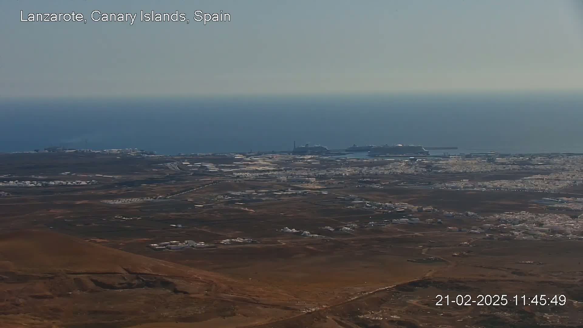 A high-angle, long shot shows a hazy, sunny day overlooking a coastal town with a port containing cruise ships, all situated next to a calm ocean.