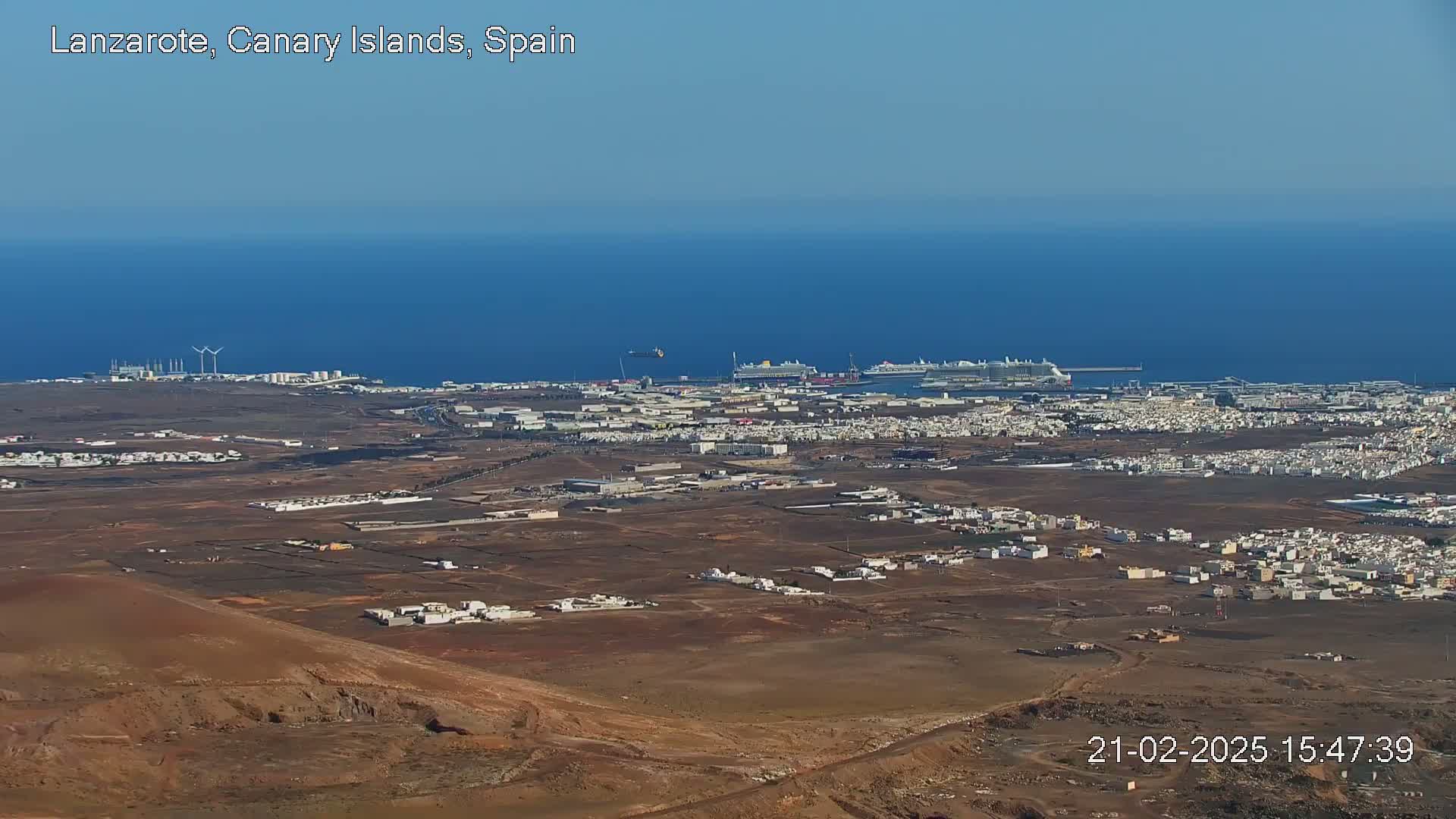 A high-angle view shows a sprawling, arid landscape with low-lying buildings and a port with several large ships, all under a clear, bright sky.