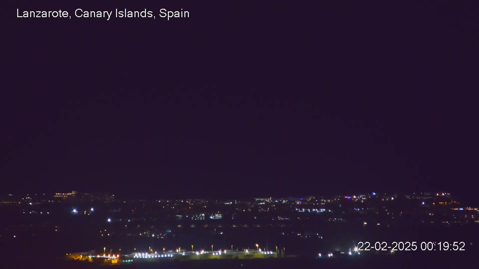 A nighttime long shot shows a city's lights spread across a dark landscape under a clear, dark sky.