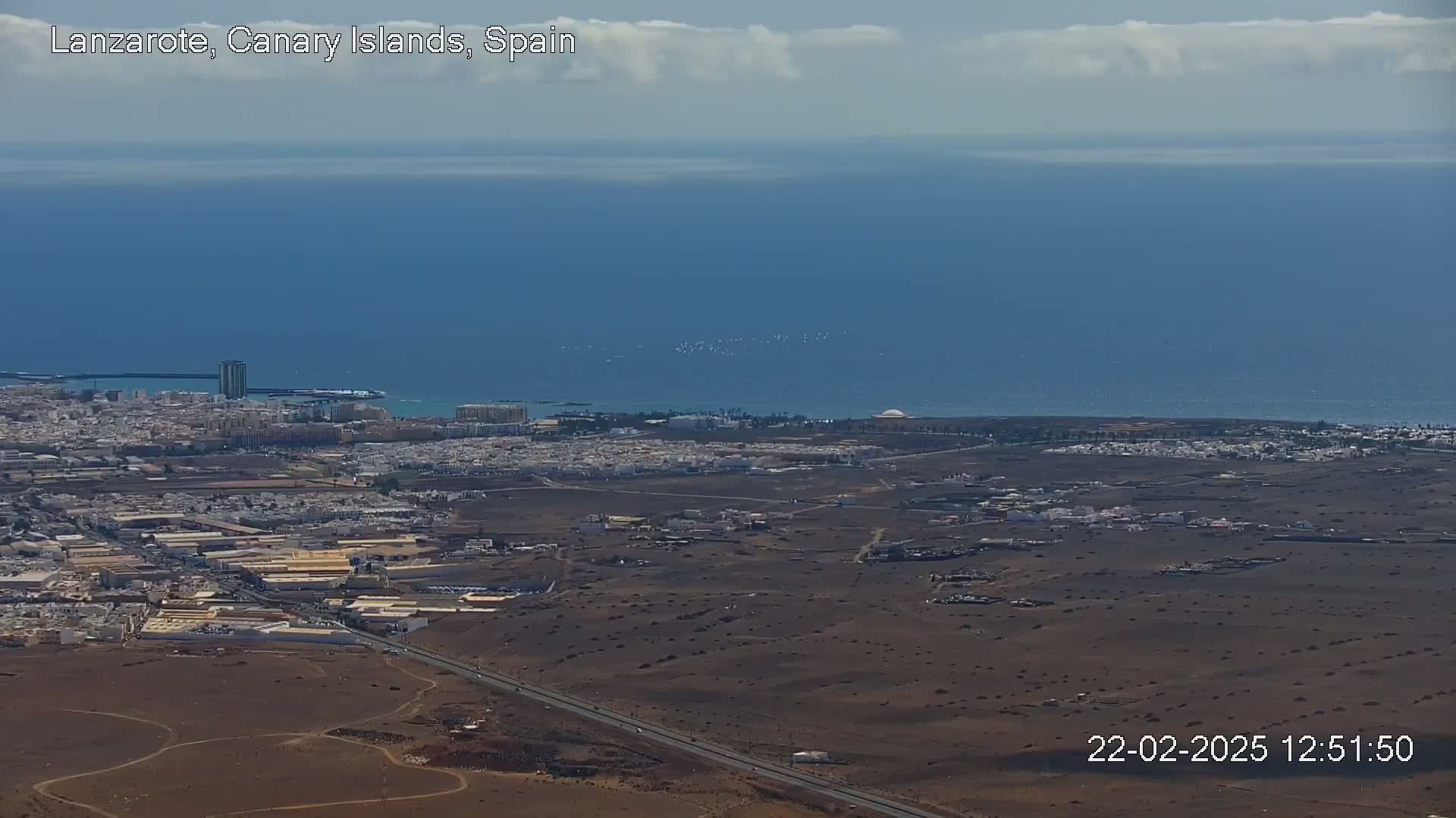 A high-angle view shows a coastal town with a mostly arid landscape, a highway, and a calm ocean under a partly cloudy sky.