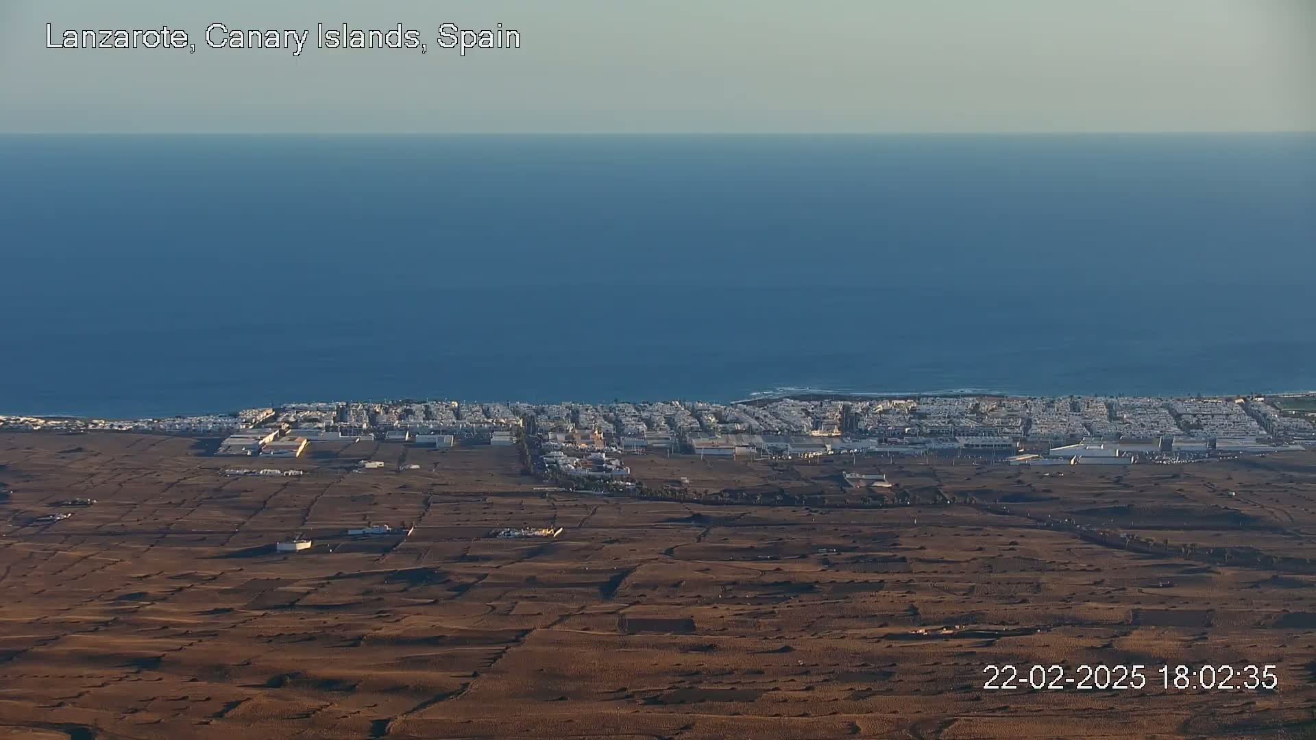 A coastal town with light brown, arid land stretches towards a calm, blue ocean under a clear sky.