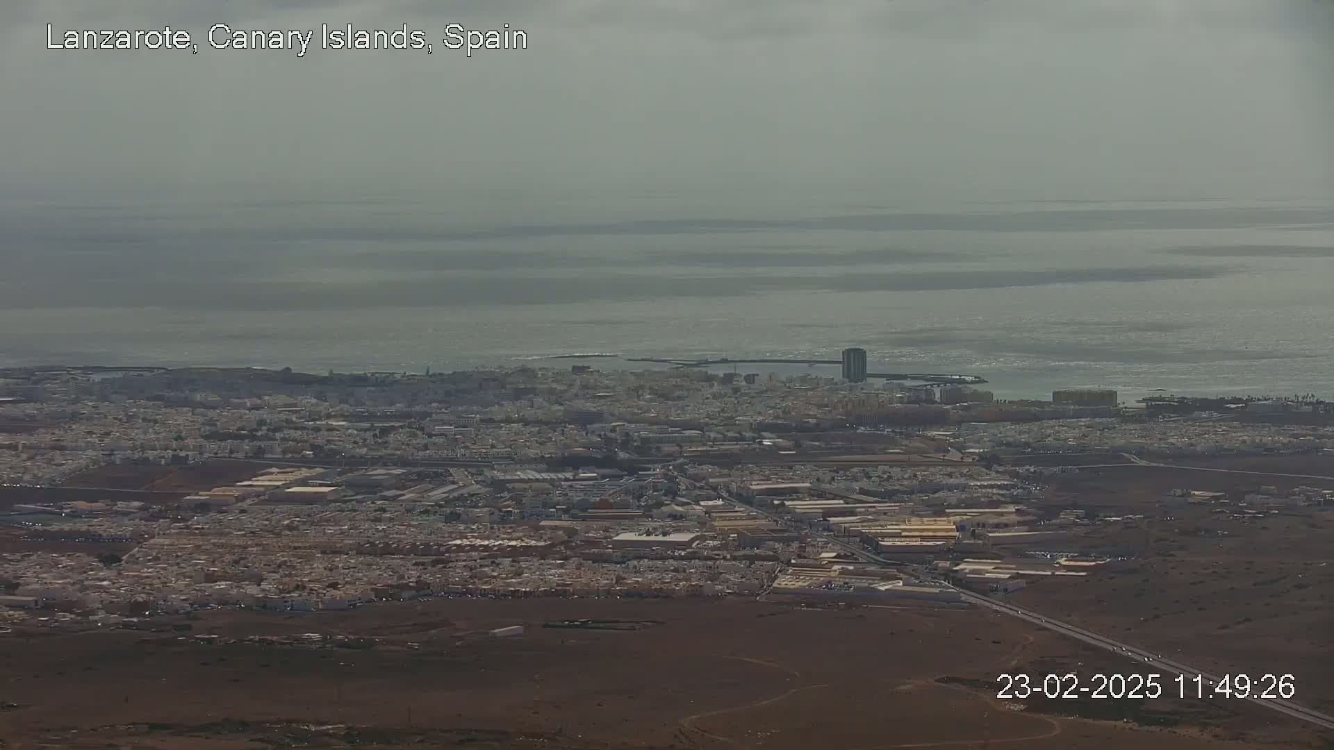 A hazy, overcast day reveals an aerial view of a coastal city sprawling across a brown landscape, adjacent to a calm, expansive body of water.