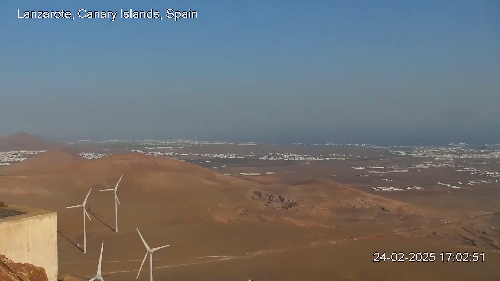 A hazy, sunny day reveals a vast, arid landscape with several wind turbines near a low wall overlooking a distant town and ocean.
