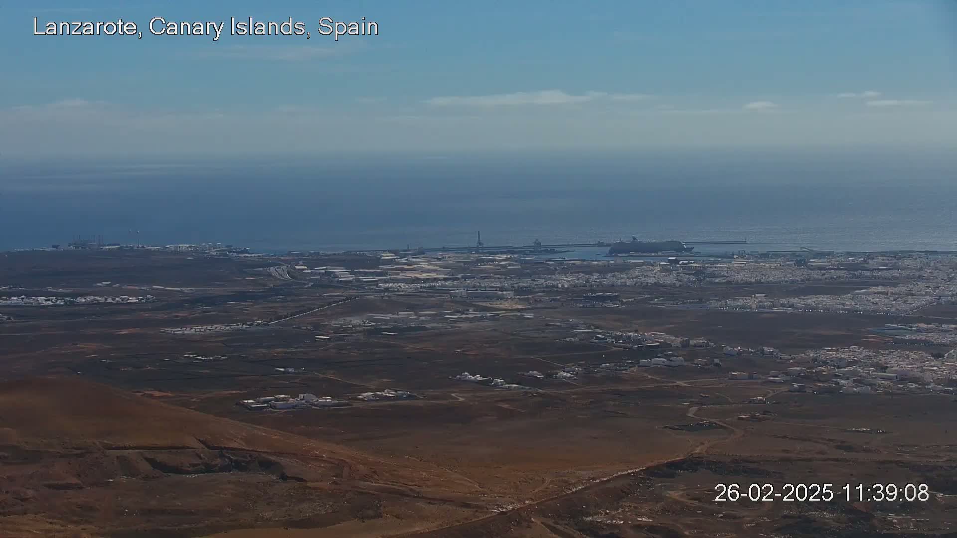 A high-angle, long shot on a clear day shows a coastal town with a port and a large ship, all set against a calm ocean.