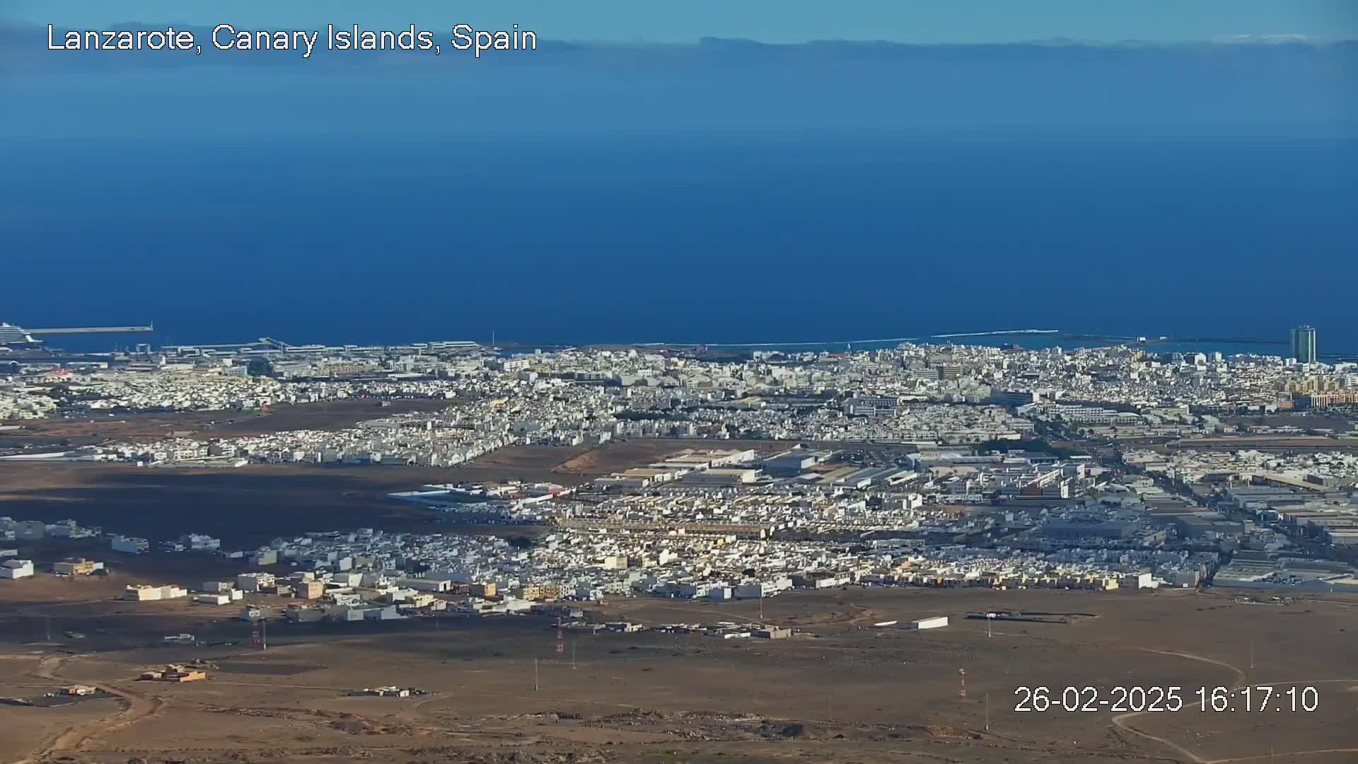 A sprawling coastal city with light-colored buildings extends to a dark-blue ocean under a clear sky.