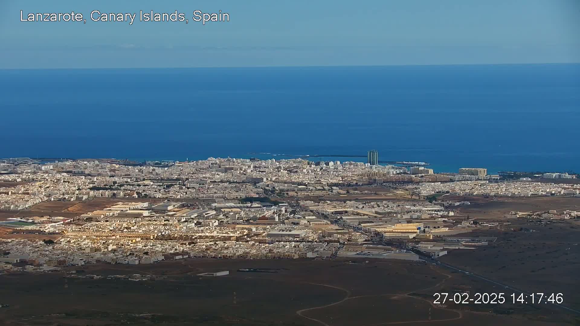 A coastal city sprawls across a largely flat landscape, meeting a calm, deep blue ocean under a clear sky.