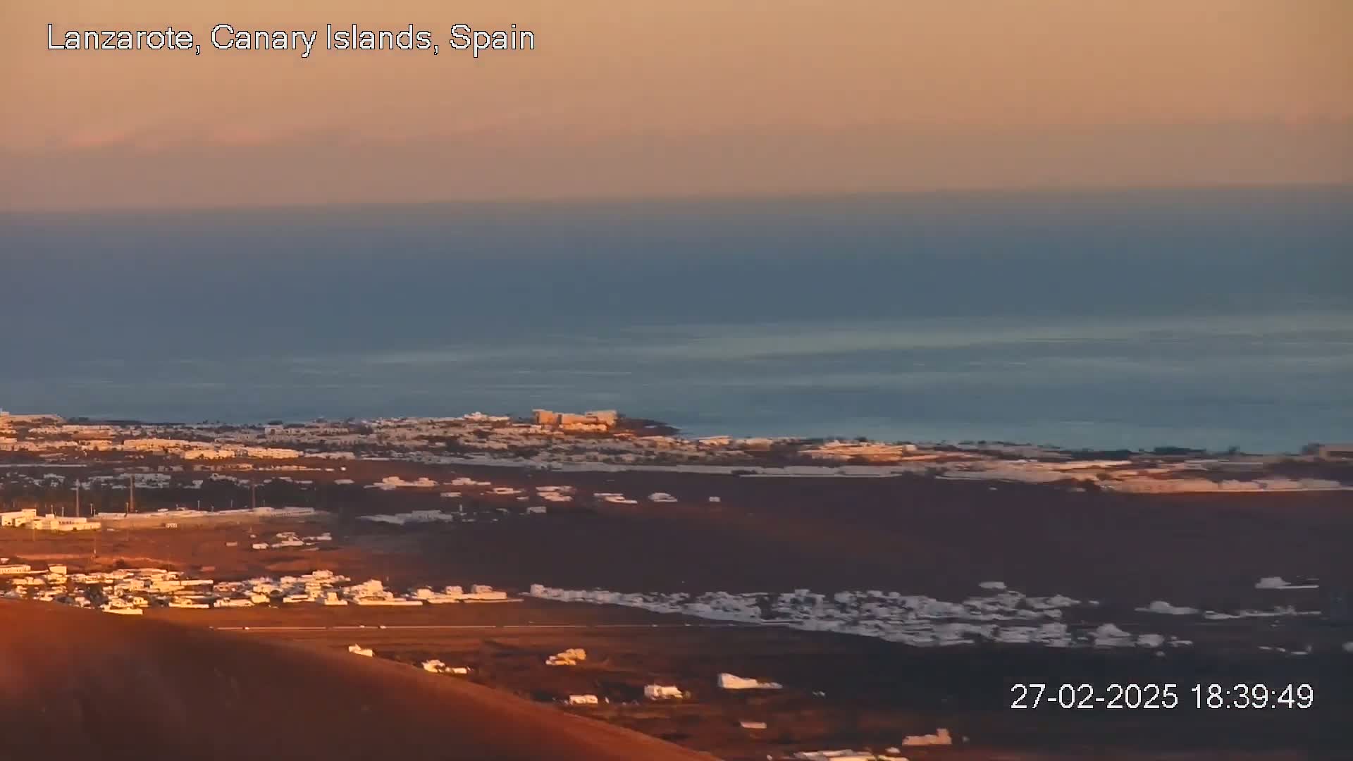 A coastal town with white buildings stretches across a volcanic landscape under a calm, sunset-colored sky.