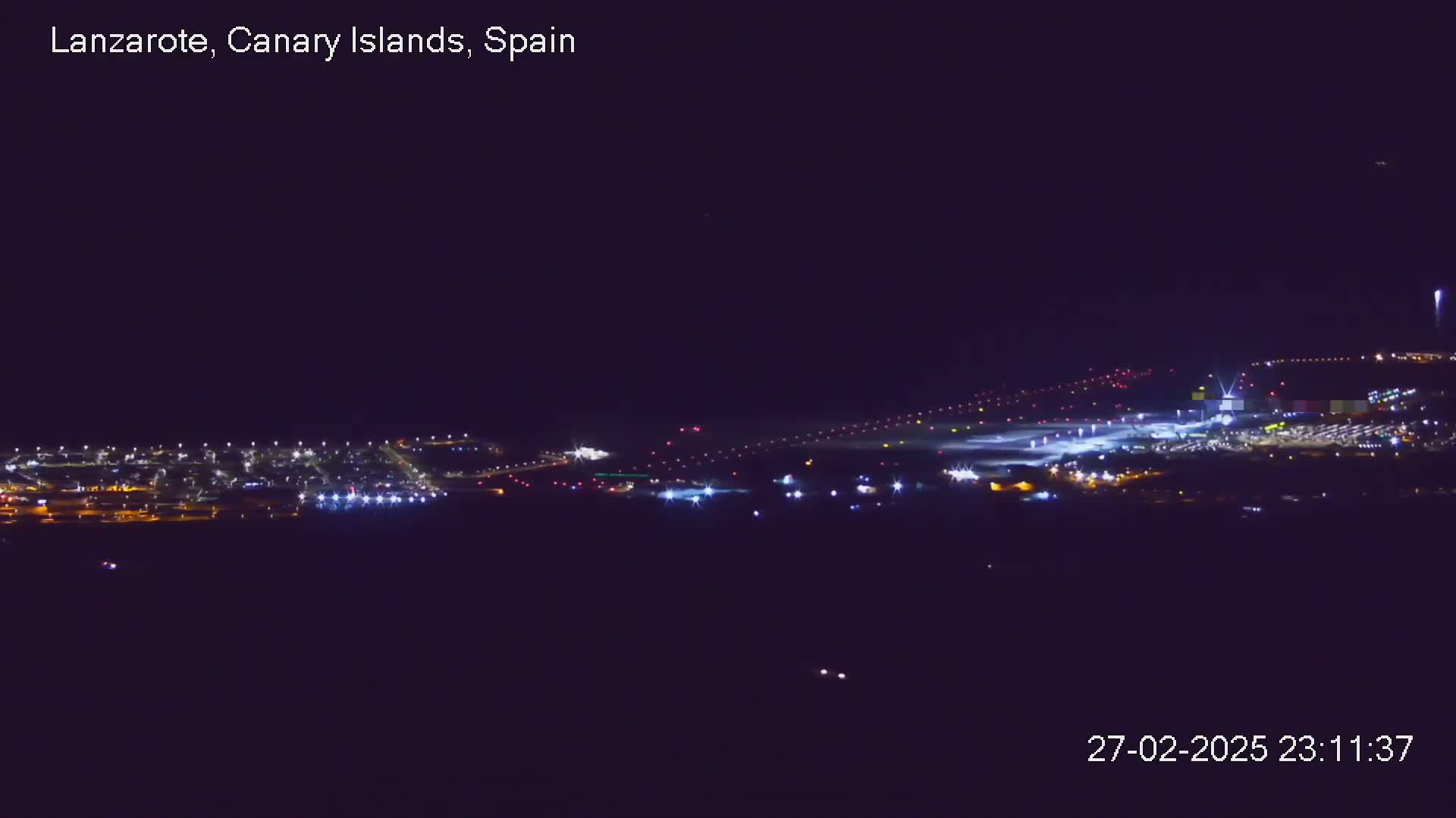 A nighttime long shot of an airport runway and surrounding buildings illuminated under a clear, dark sky.