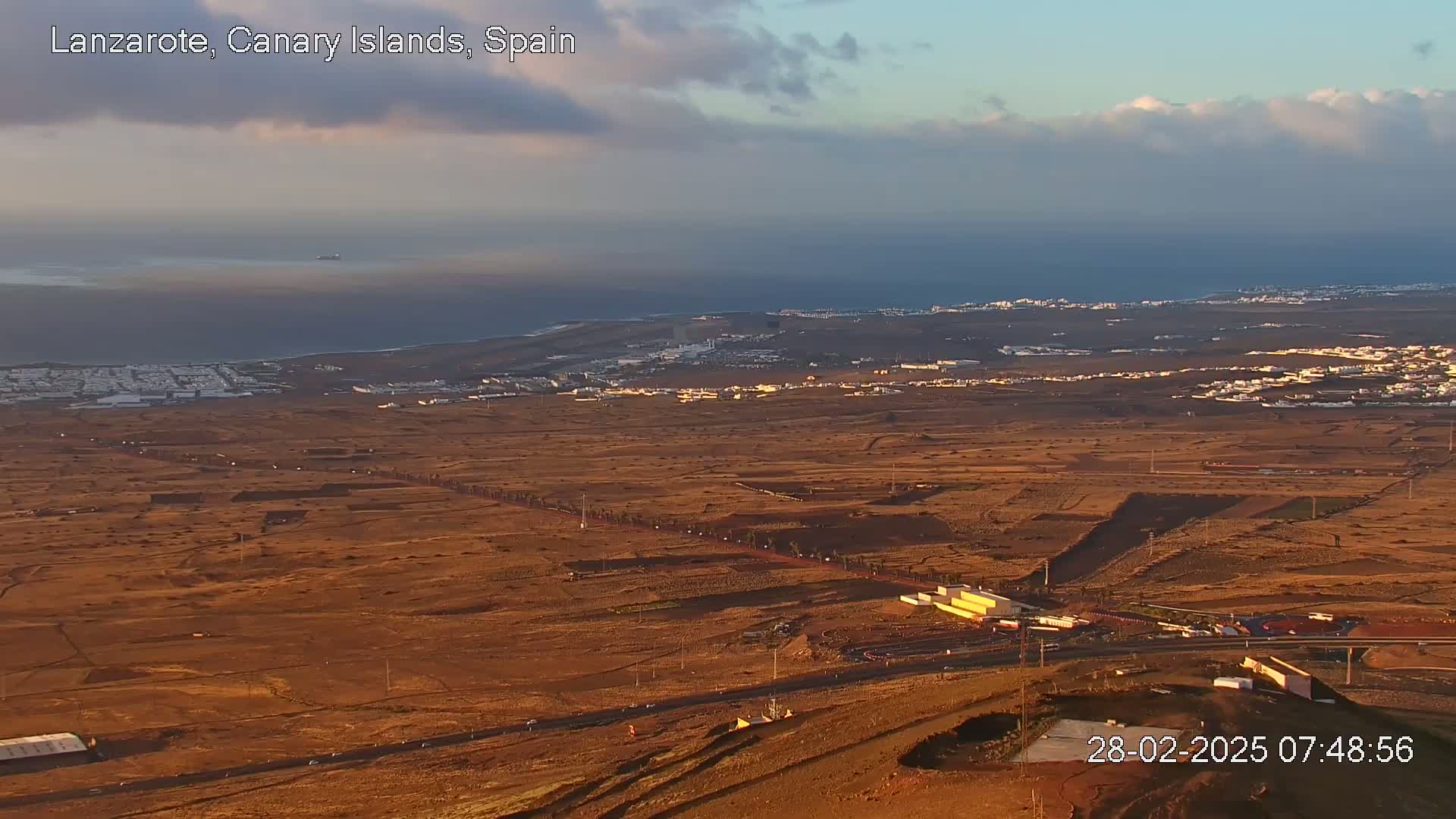 A high-angle, long shot shows a mostly arid, reddish-brown landscape with scattered buildings and roads, overlooking a calm ocean under a partly cloudy sky.