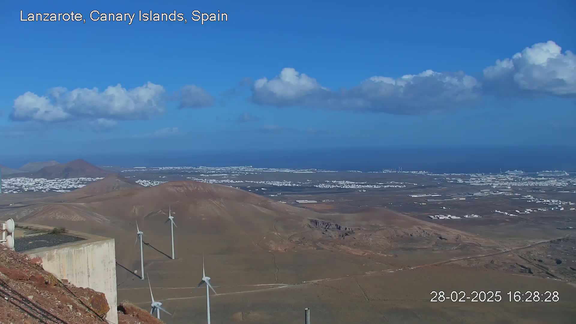 A panoramic view shows a volcanic landscape with wind turbines, a coastal town, and a partly cloudy blue sky.