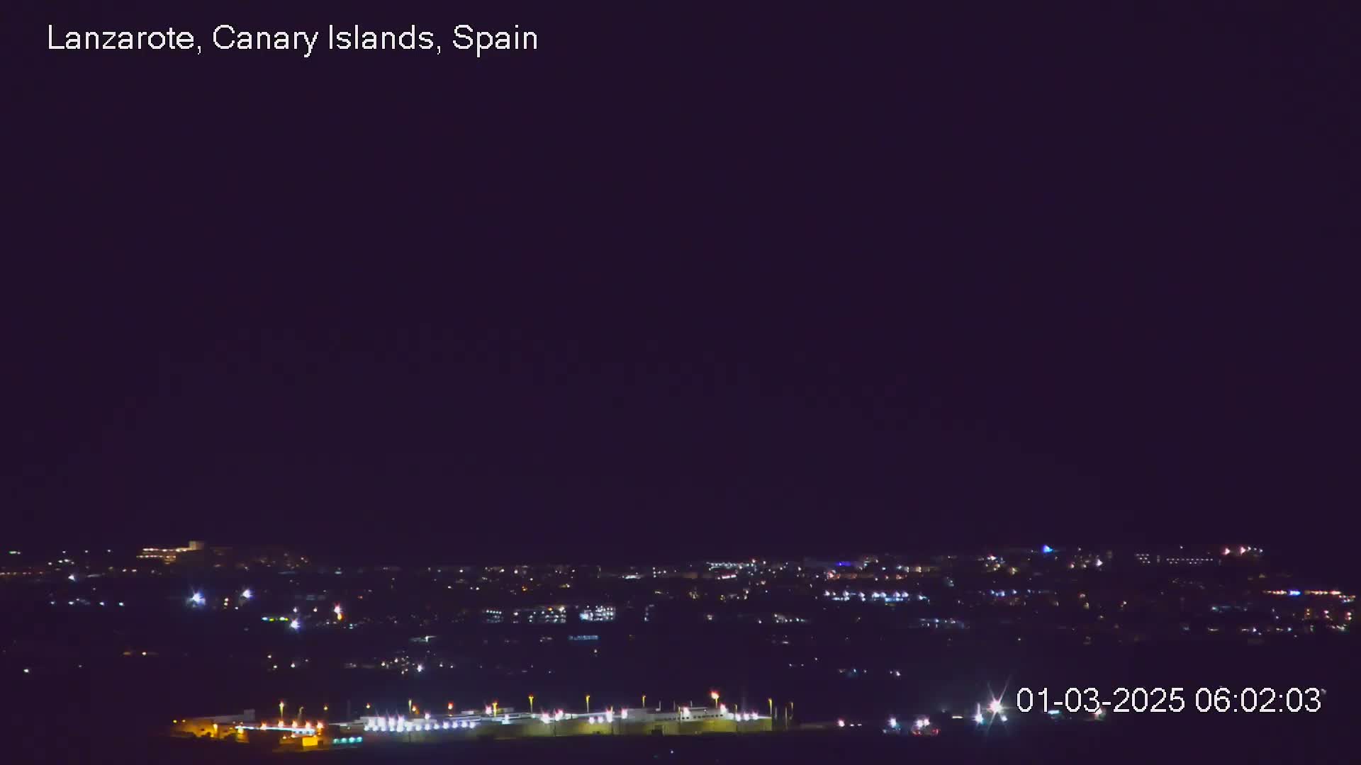 A nighttime long shot shows a city's lights spread across a dark landscape under a clear, dark sky.