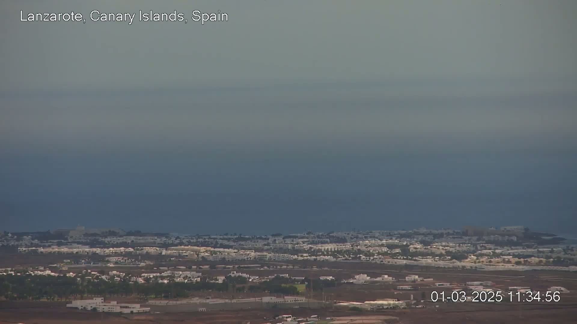 A hazy, overcast view shows a coastal town with low-lying white buildings stretching to a dark blue ocean under a muted sky.