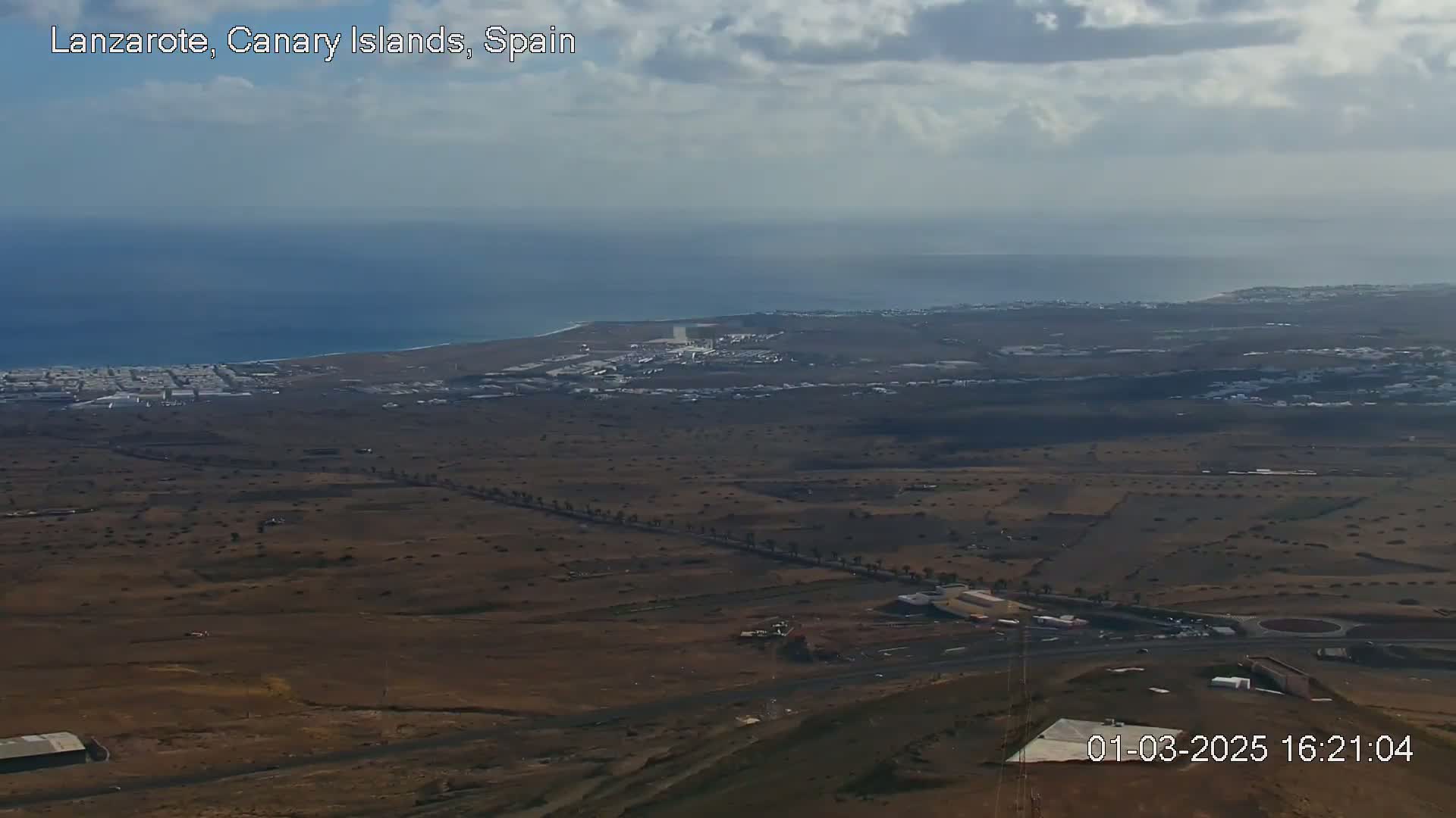 A high-angle, long shot shows a mostly-brown, arid landscape with a coastline and ocean in the background under a partly cloudy sky.