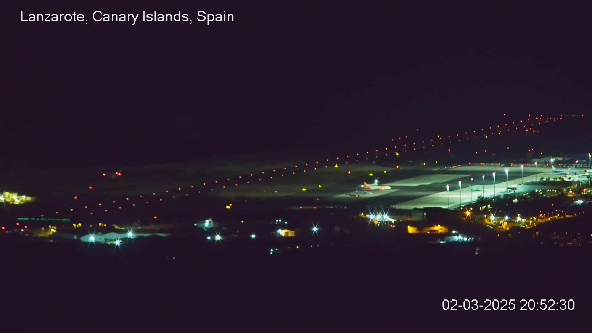 A brightly lit airport runway at night, with a plane visible on the tarmac, is seen from a distance under a clear, dark sky.