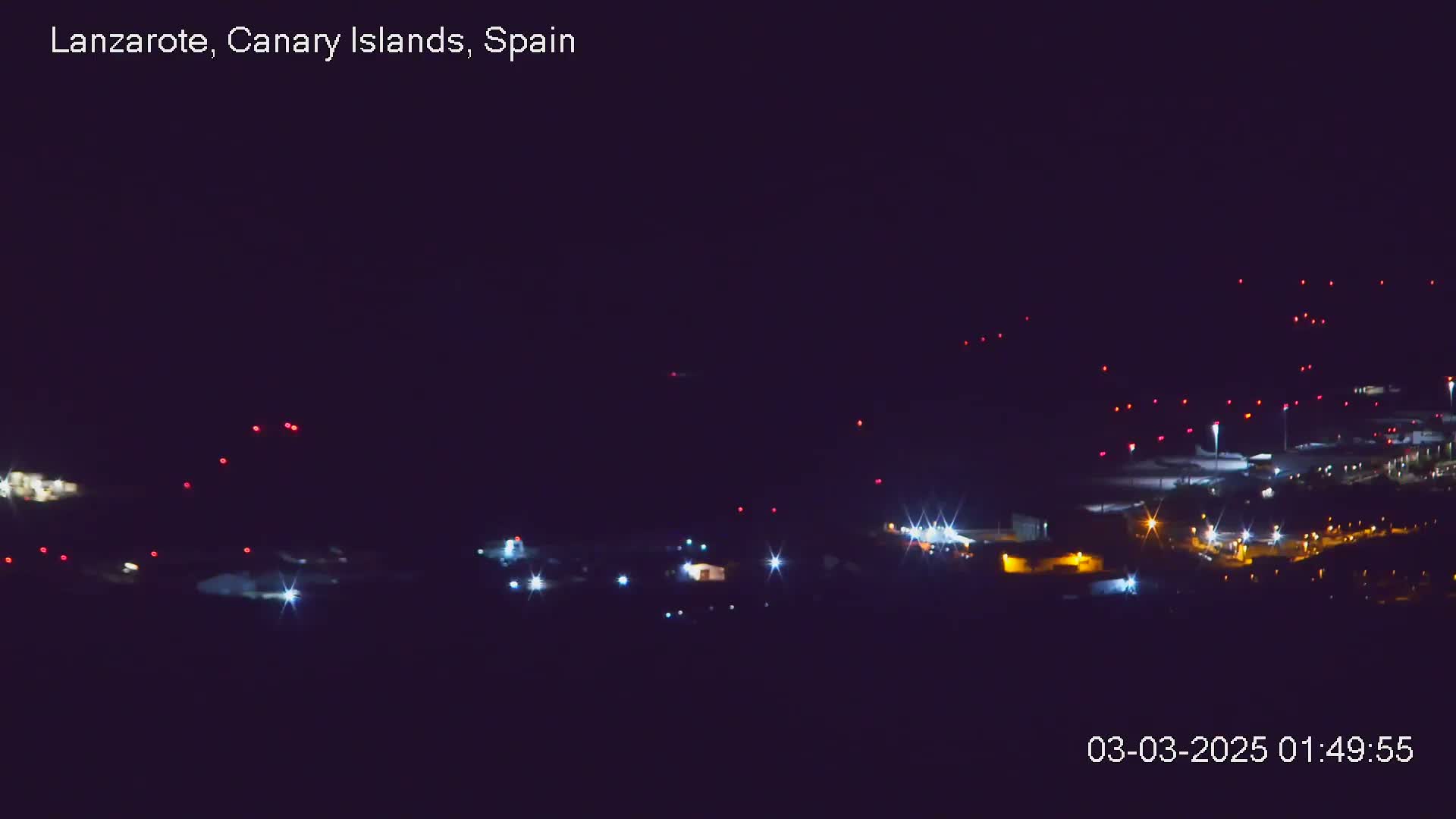 A nighttime long shot of an airport with numerous red and white lights illuminating the tarmac and buildings under a clear, dark sky.