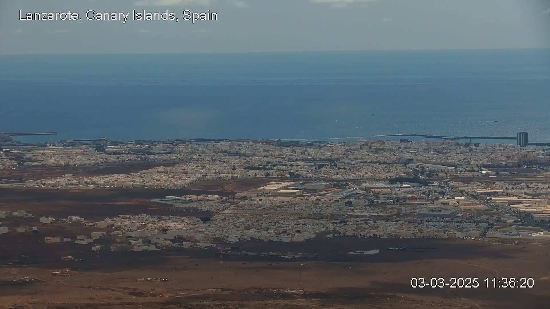 A sprawling coastal city with light-colored buildings stretches towards a calm, blue ocean under a mostly clear sky.