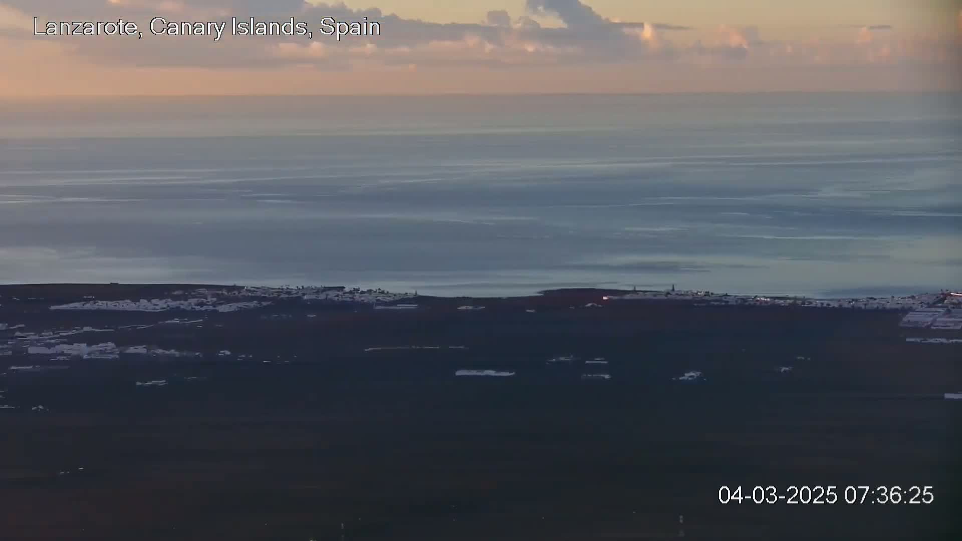 A coastal town with white buildings is visible at the bottom of a wide expanse of calm ocean under a mostly clear, dusky sky.