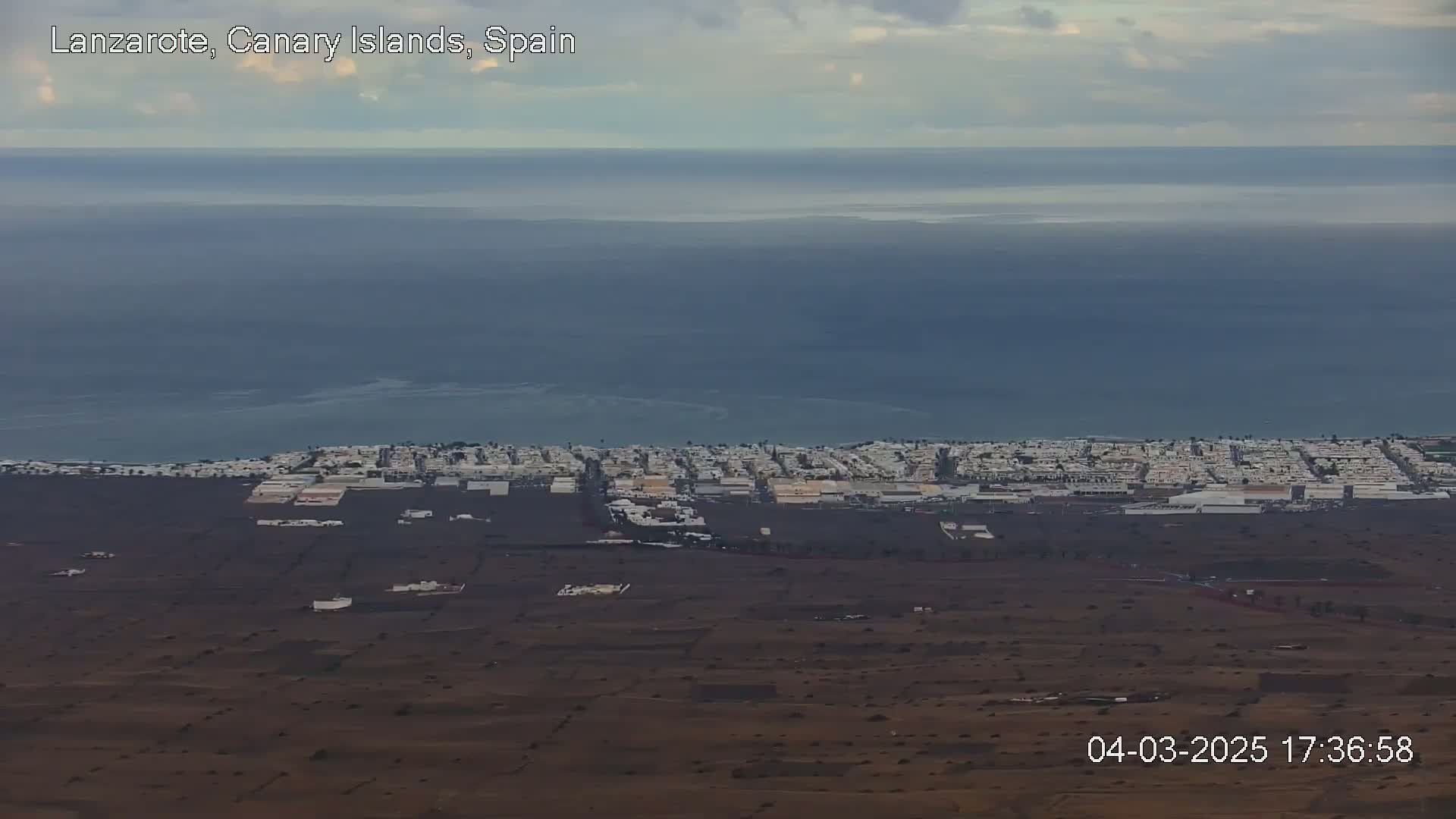 A coastal town with light-colored buildings stretches along a dark-colored shoreline, overlooking a calm, blue ocean under a mostly cloudy sky.