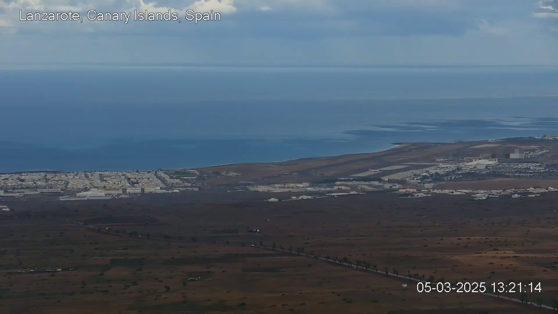 A mostly clear day reveals a coastal town near an airport, extending to a calm ocean under a slightly cloudy sky.
