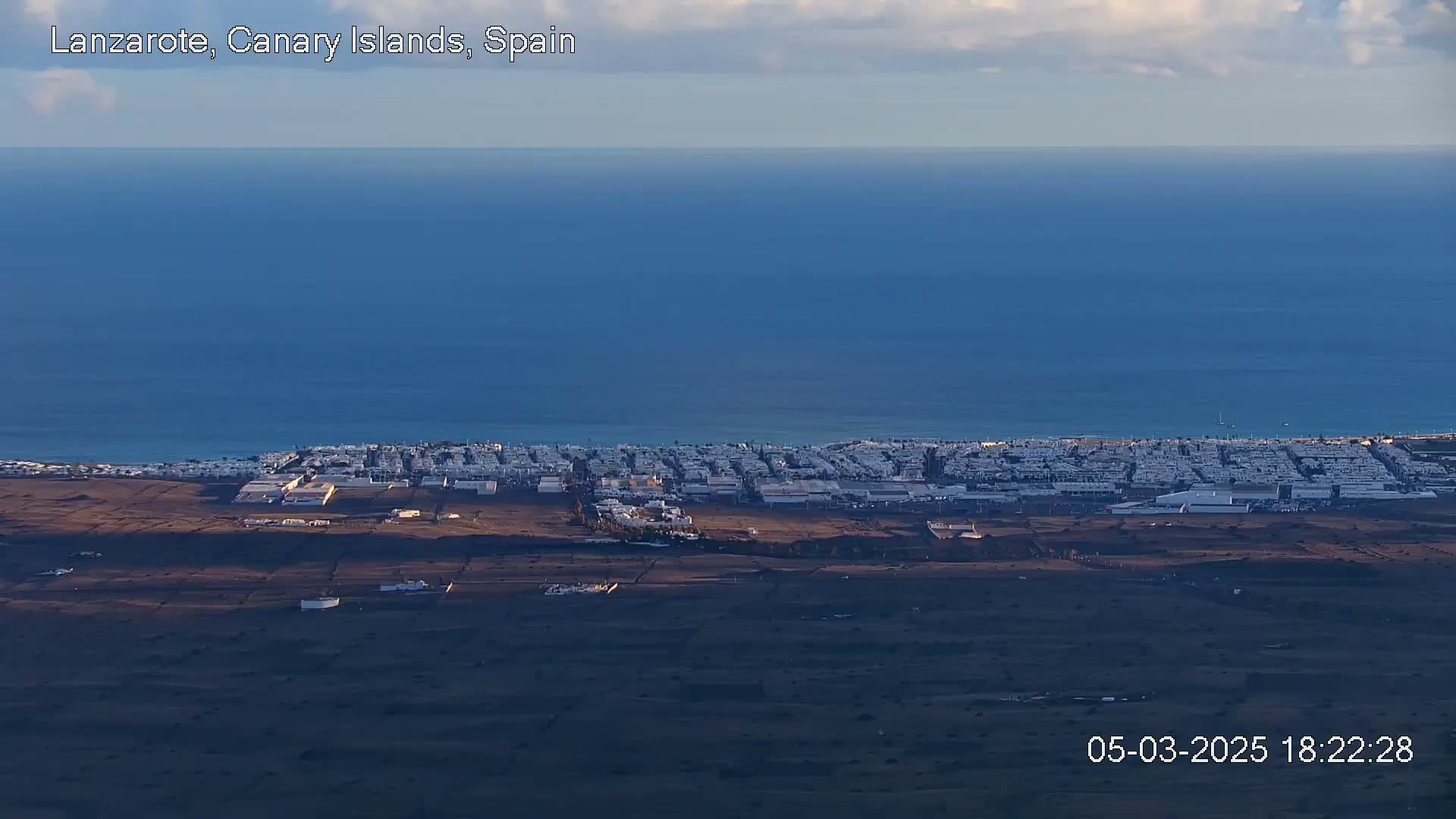 A coastal town with white buildings stretches along a dark-blue ocean under a mostly clear sky.