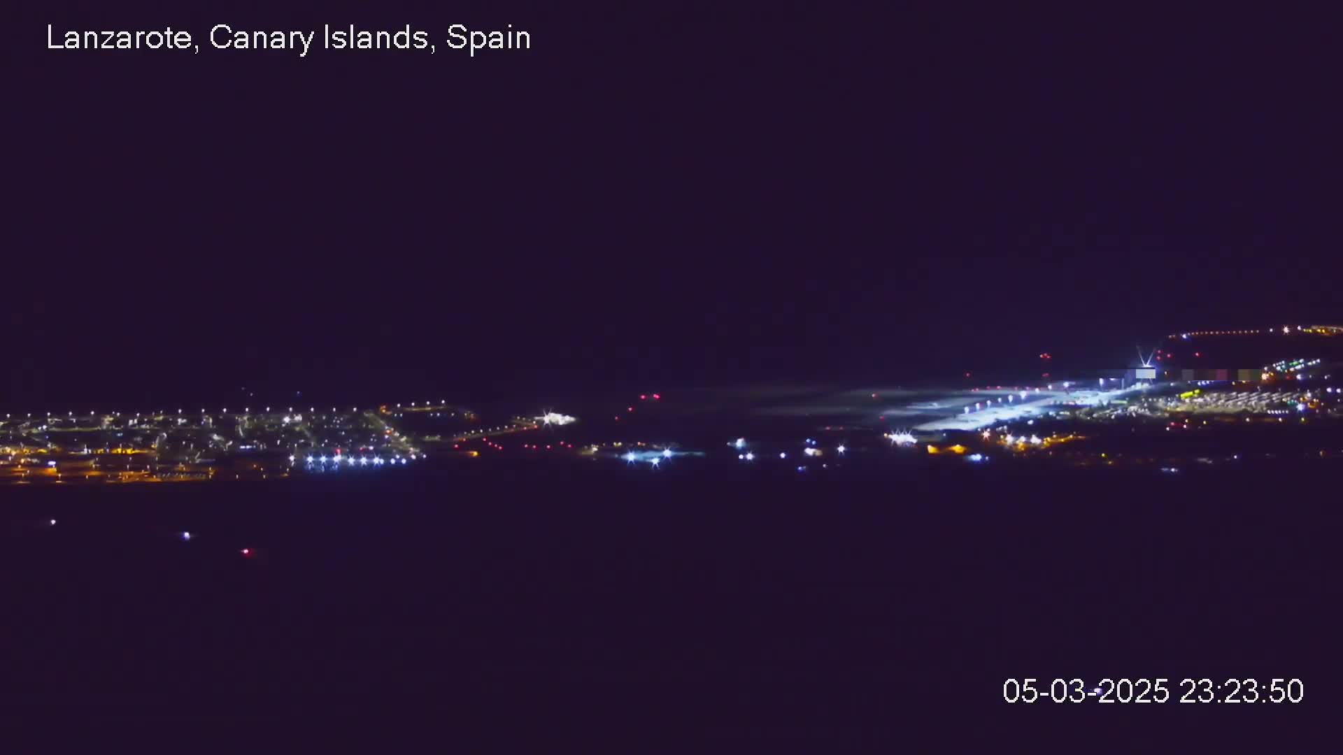 A brightly lit airport is visible at night, with some light fog or haze obscuring the distance.