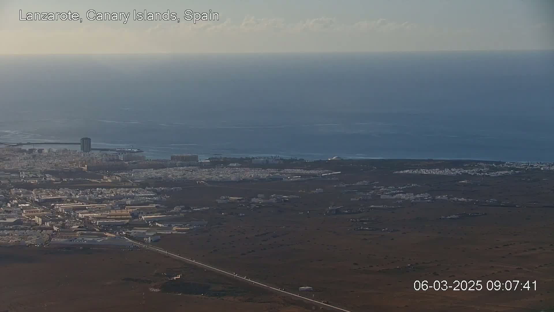 A coastal town with white buildings stretches across a dark brown, volcanic landscape next to a calm, blue ocean under a mostly clear sky.
