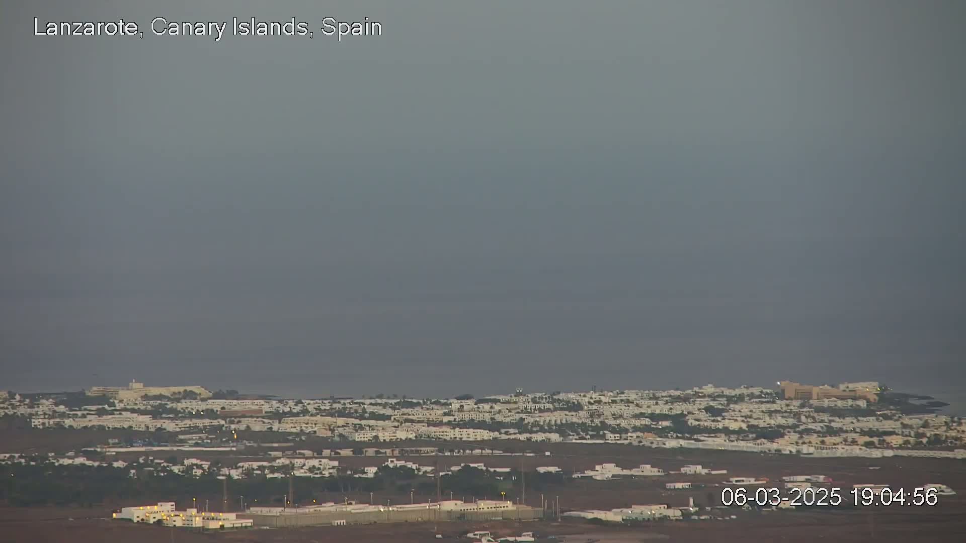 A hazy, twilight view shows a coastal town with low-rise white buildings, extending to a calm ocean under a muted sky.