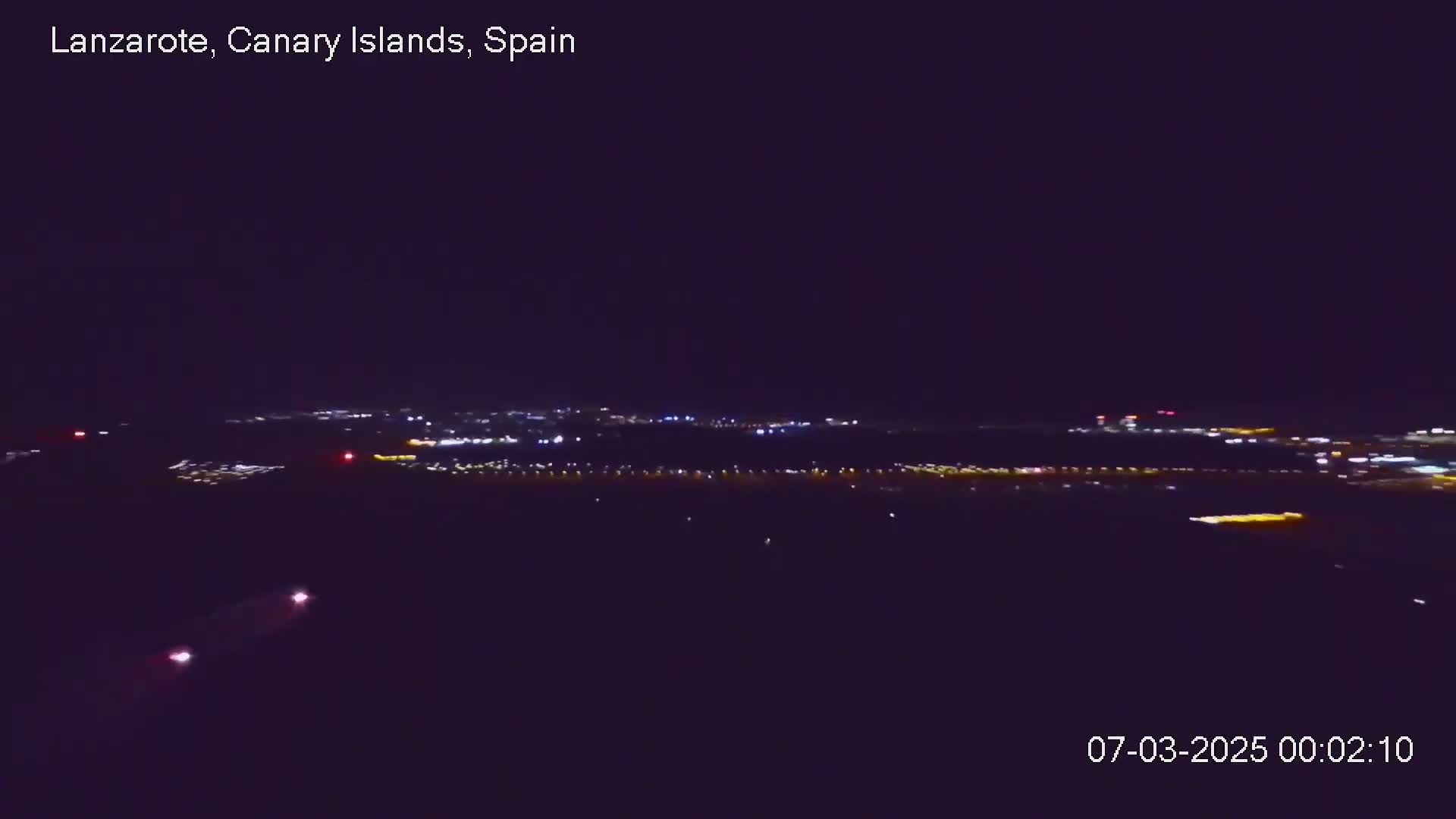 A nighttime long shot shows a distant city illuminated by lights under a dark, clear sky.