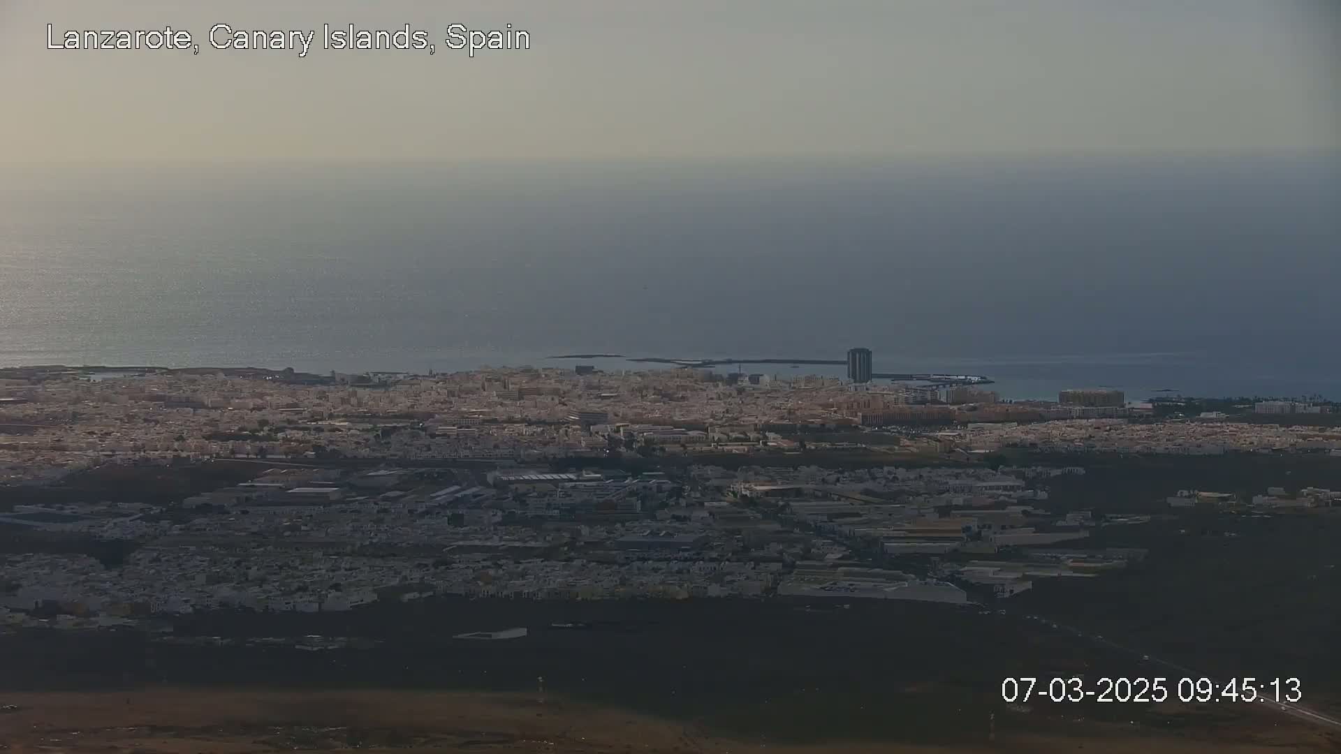 An expansive view of a coastal city with a hazy sky overlooks a dark volcanic landscape under overcast conditions.