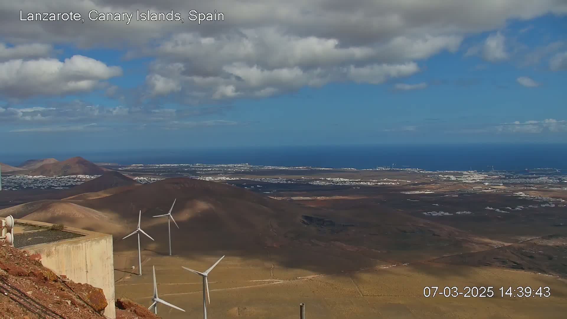 A partly cloudy daytime view of a volcanic landscape with wind turbines, overlooking a coastal town and ocean.