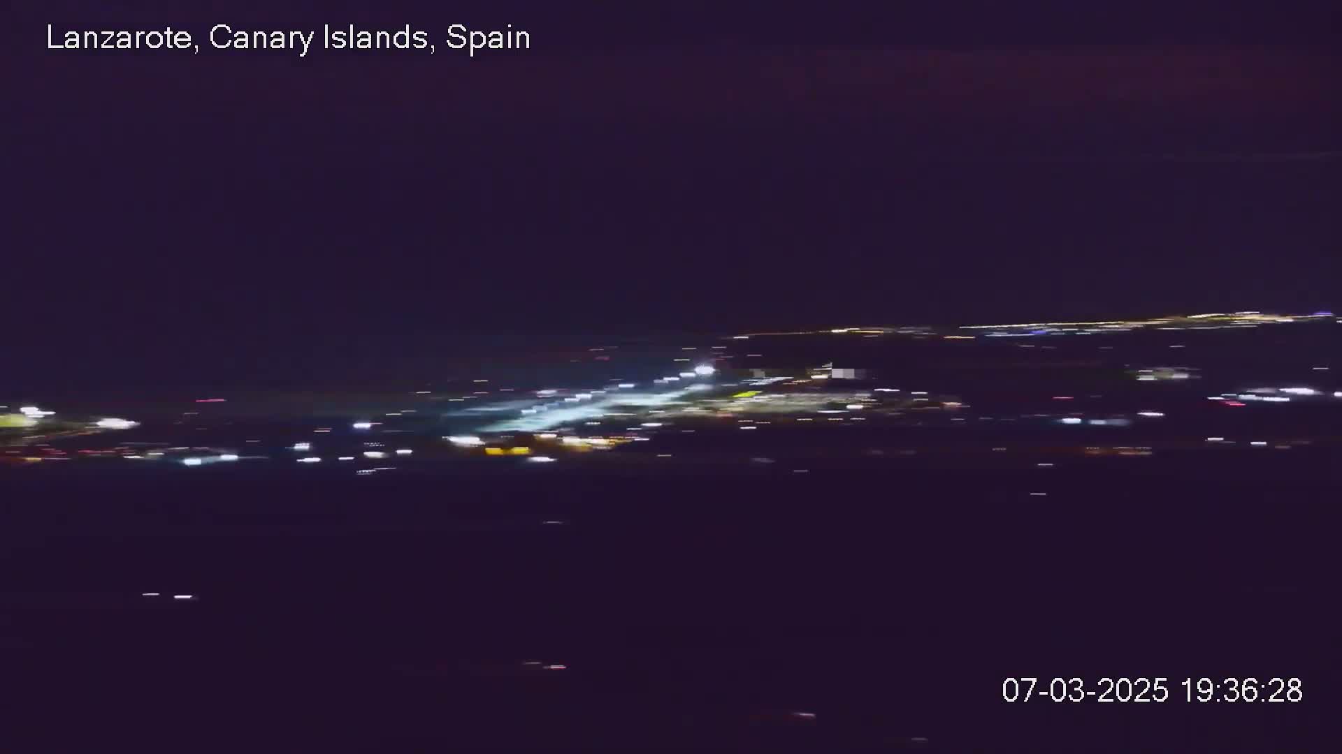 A nighttime aerial view shows a brightly lit airport runway and surrounding area under a dark, clear sky.