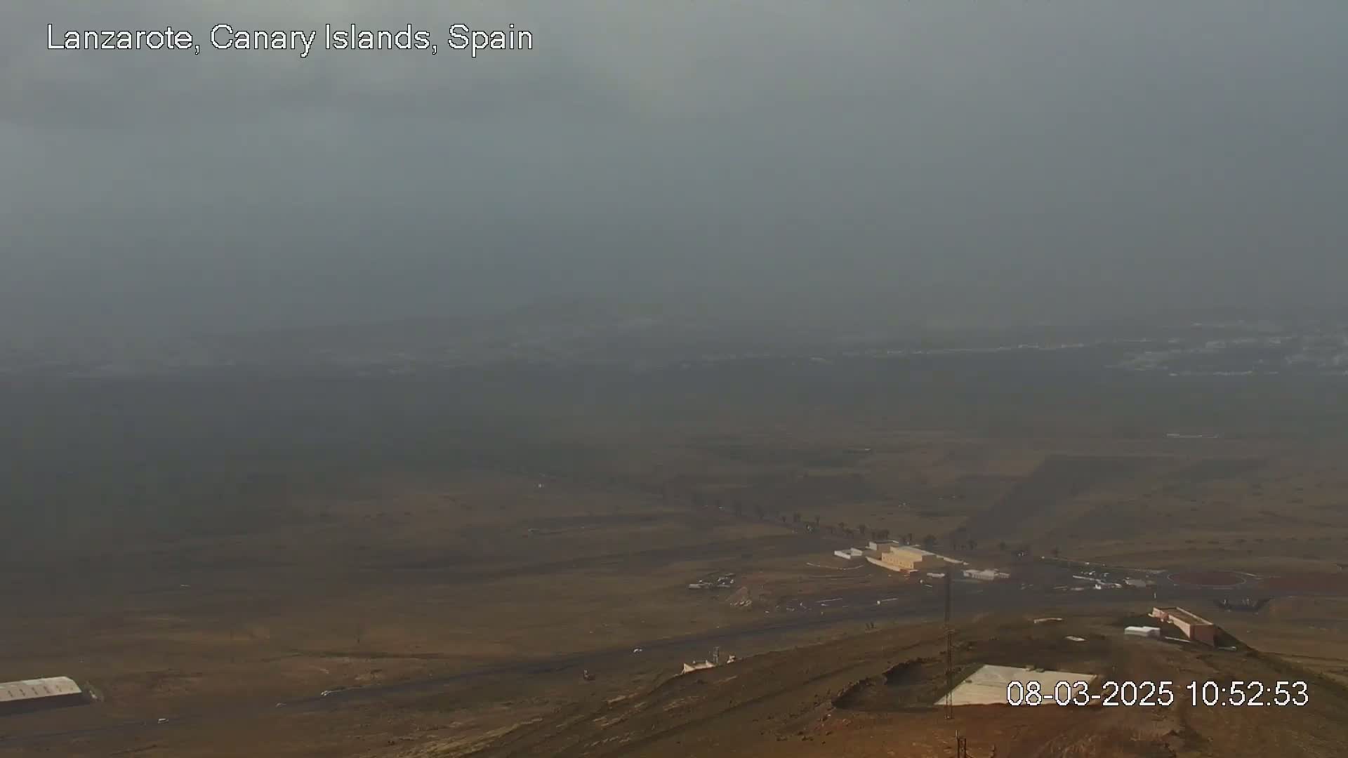 A hazy, distant view of a flat, brown landscape with a few structures, possibly a parking lot and buildings, under a gray sky.