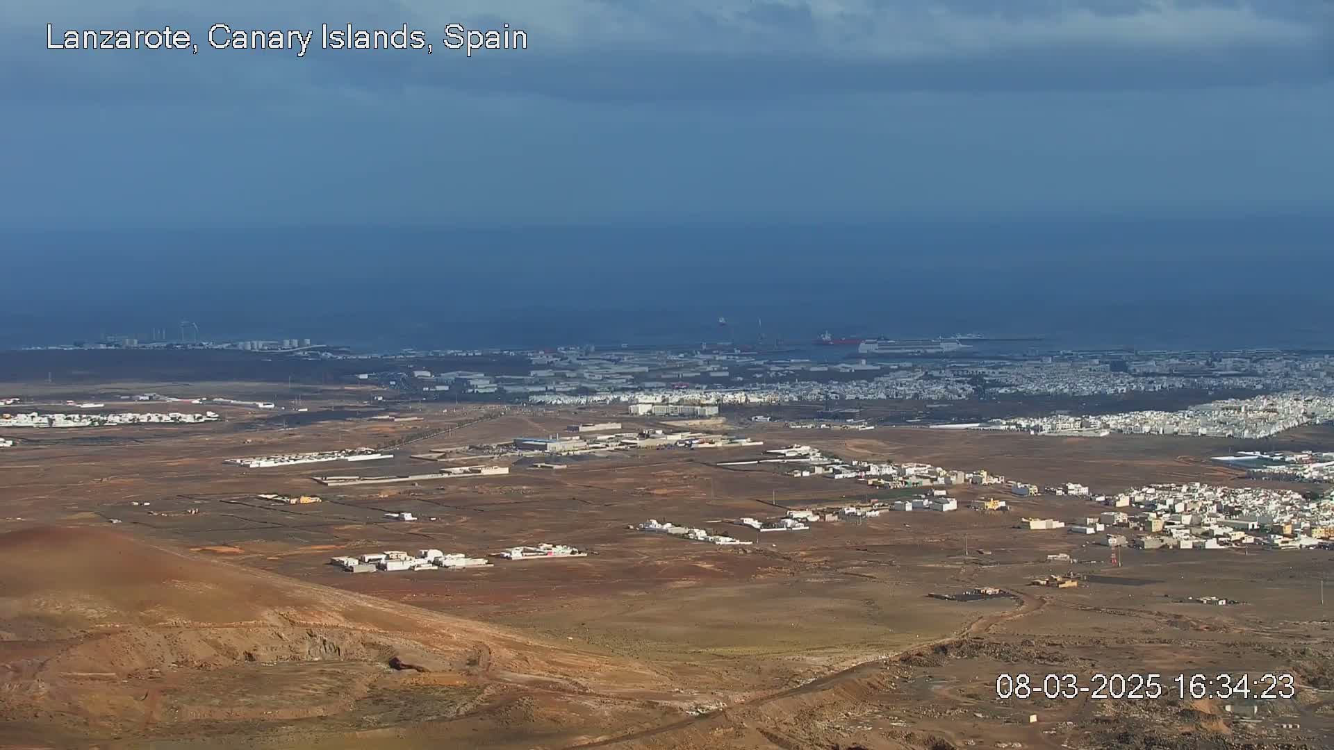 A high-angle view shows a mostly sunny, dry landscape with a town and industrial areas near the ocean under a partly cloudy sky.