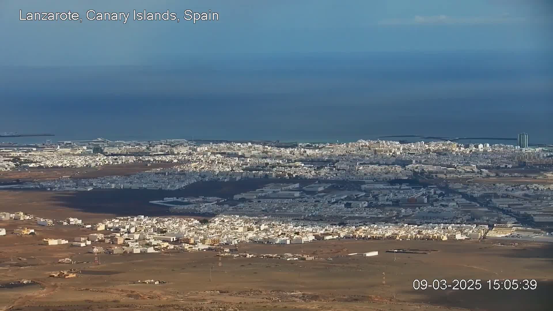 A sprawling city with white buildings extends to a calm blue ocean under a clear sky.