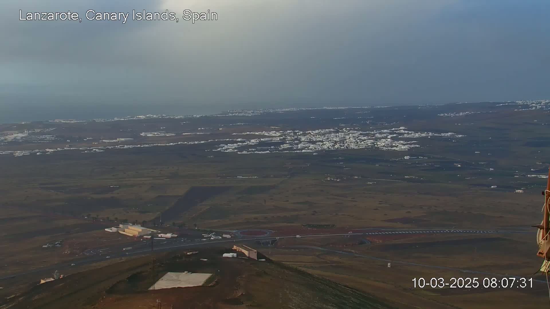 A high-angle, long shot shows a hazy, overcast day overlooking a coastal town and surrounding flatlands, with a portion of an airport visible in the foreground.