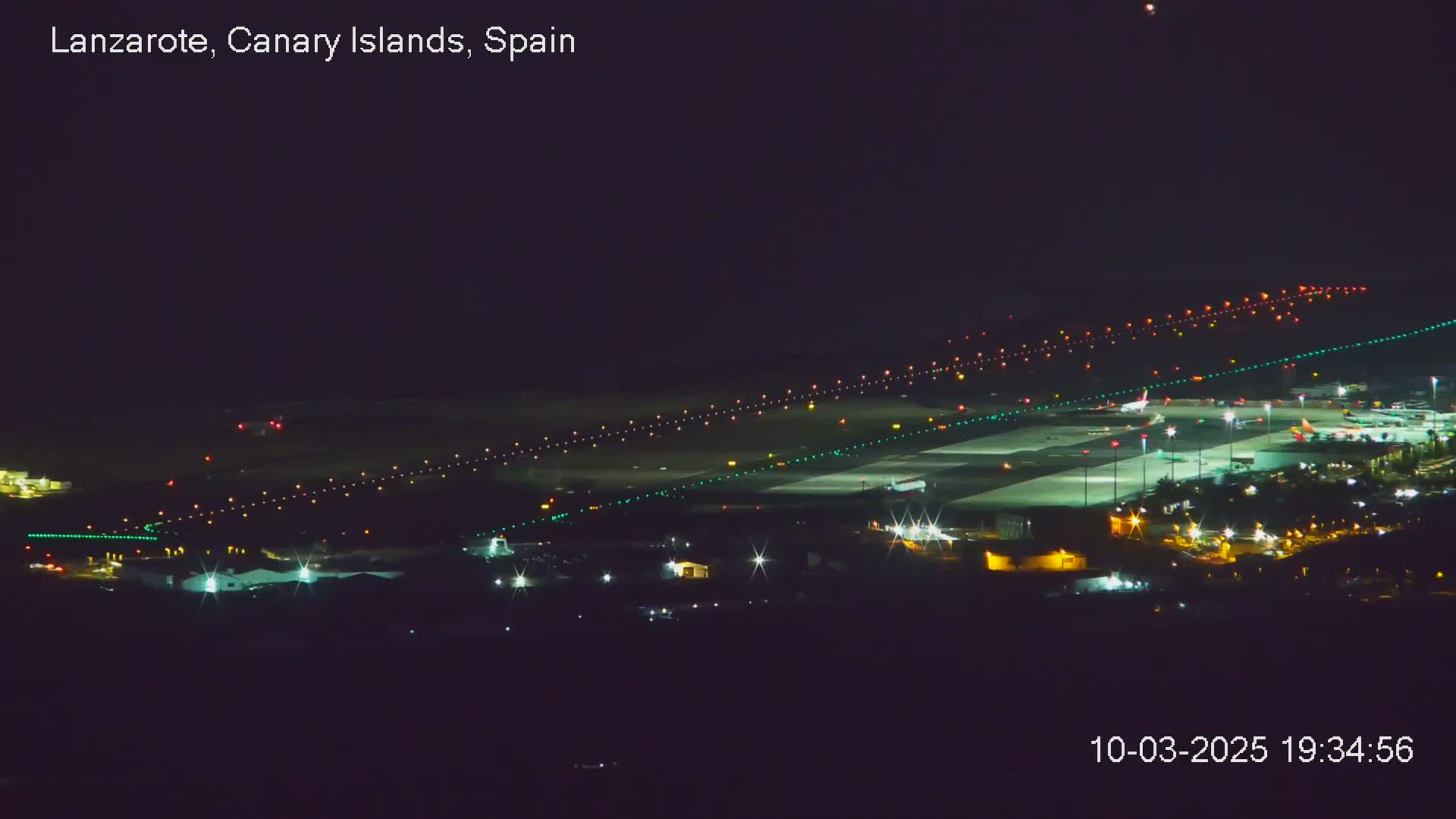 A brightly lit airport runway and surrounding buildings are visible at night under a clear, dark sky.
