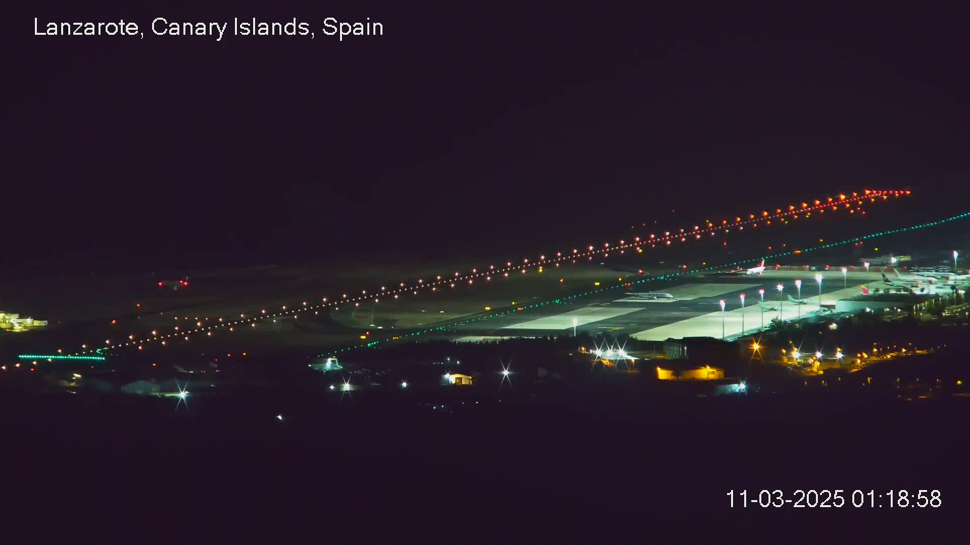 A brightly lit airport runway and tarmac at night, with several airplanes parked and one possibly taxiing.