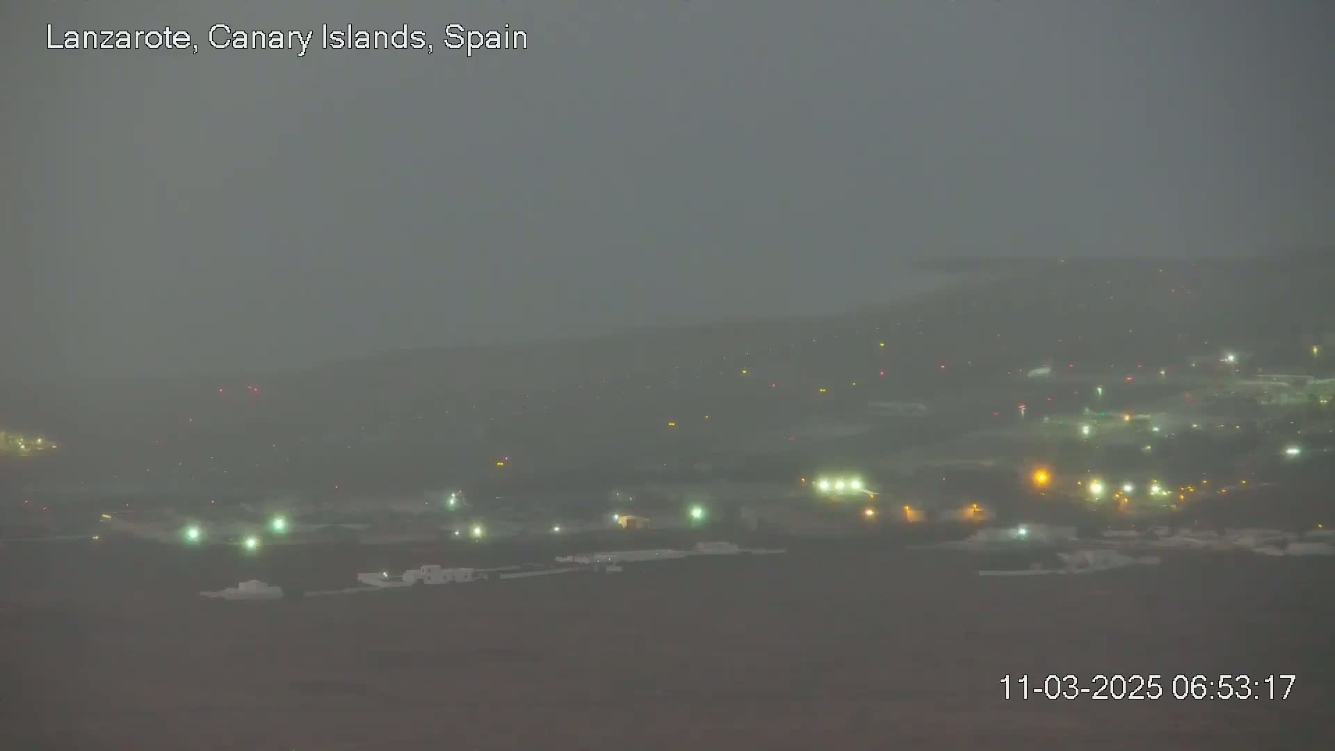 A hazy, dimly lit landscape at night shows a collection of buildings and lights in the distance under a foggy sky.