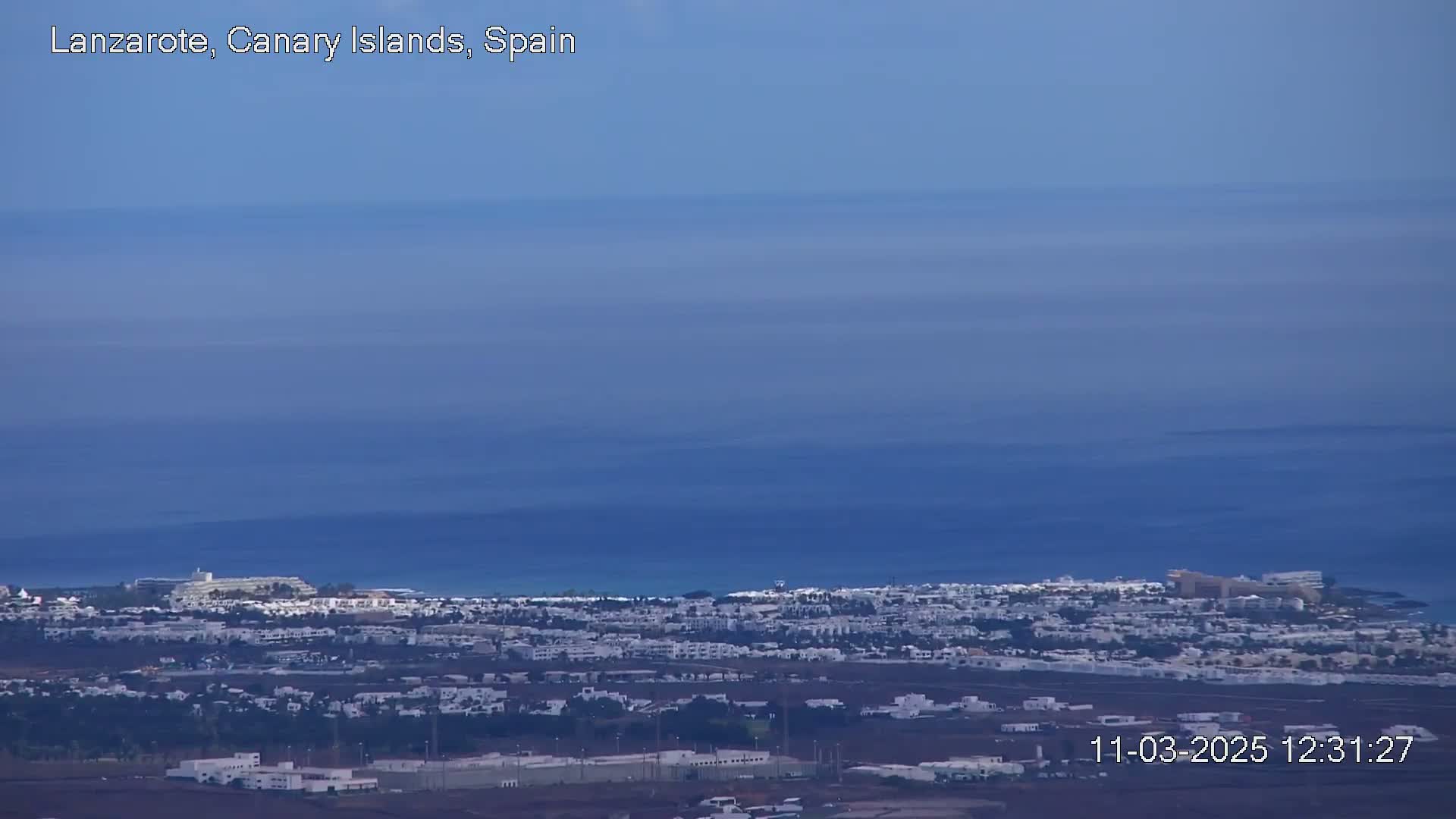 A coastal town with white buildings stretches along a calm, blue ocean under a clear sky.