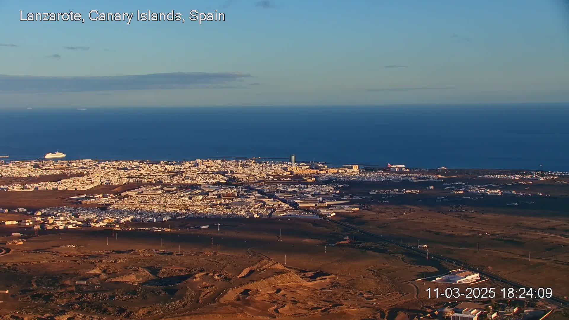 A coastal city with a large expanse of dark-brown desert landscape is seen from an aerial view on a clear, sunny day, with a calm blue ocean in the background.