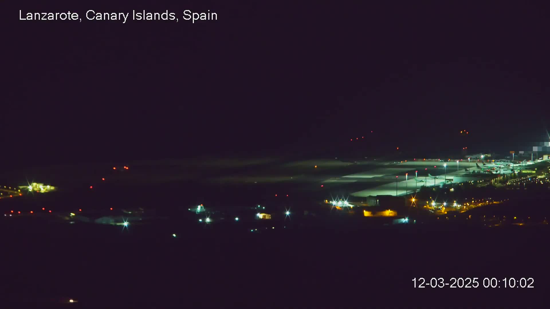 A nighttime long shot of an airport with planes on the tarmac, illuminated runways, and surrounding buildings.