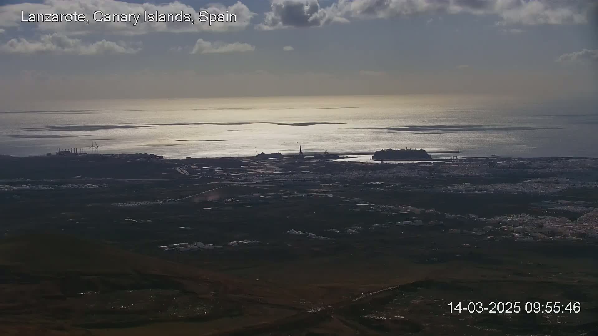 A high-angle, long shot shows a coastal town and harbor under a partly cloudy sky, with a calm sea reflecting sunlight.