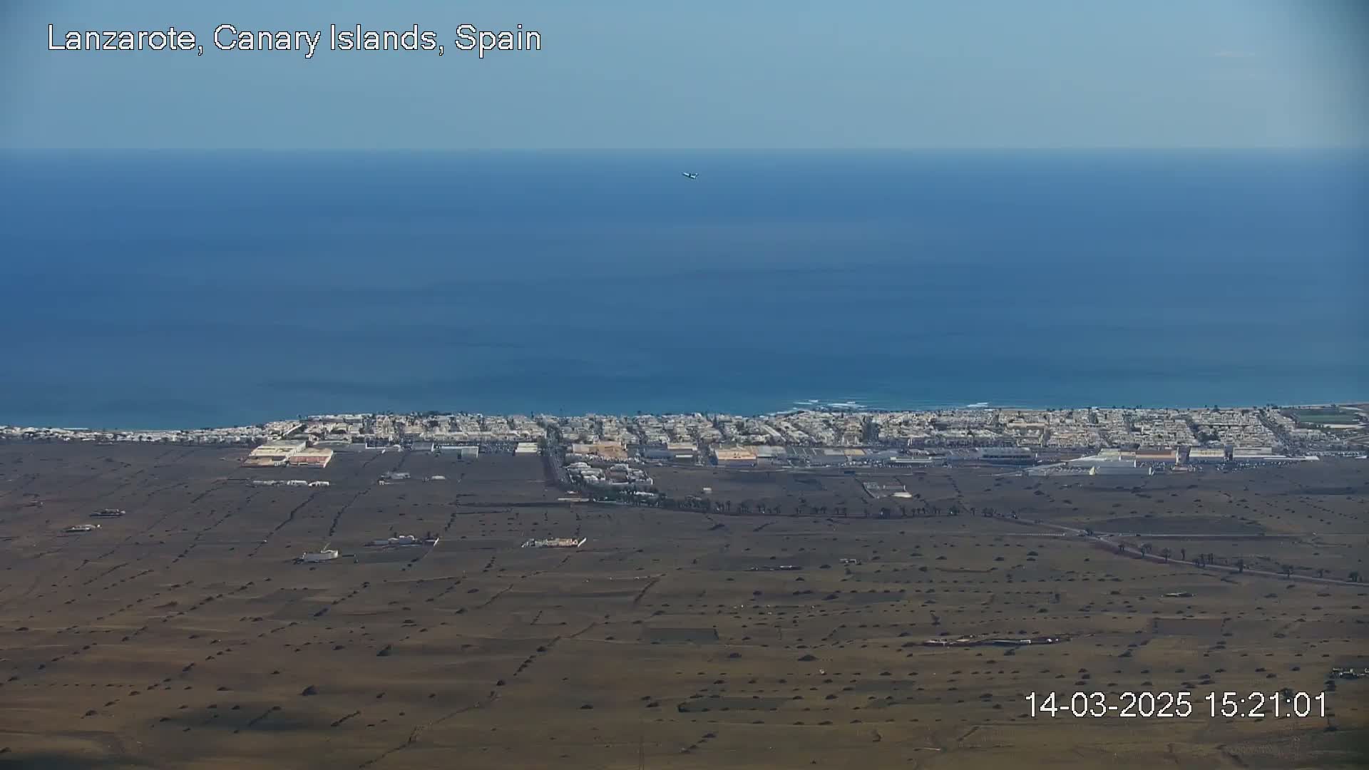 A coastal town is visible below a clear blue sky and ocean, with brown, sparsely vegetated land separating the town from the viewer's perspective.