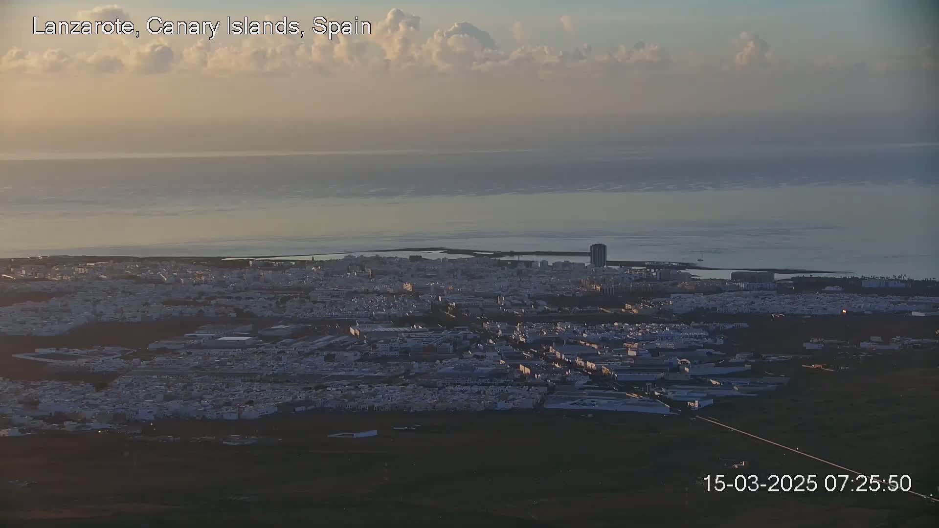 A coastal city with white buildings extends to a calm ocean under a mostly cloudy, pale sunrise sky.