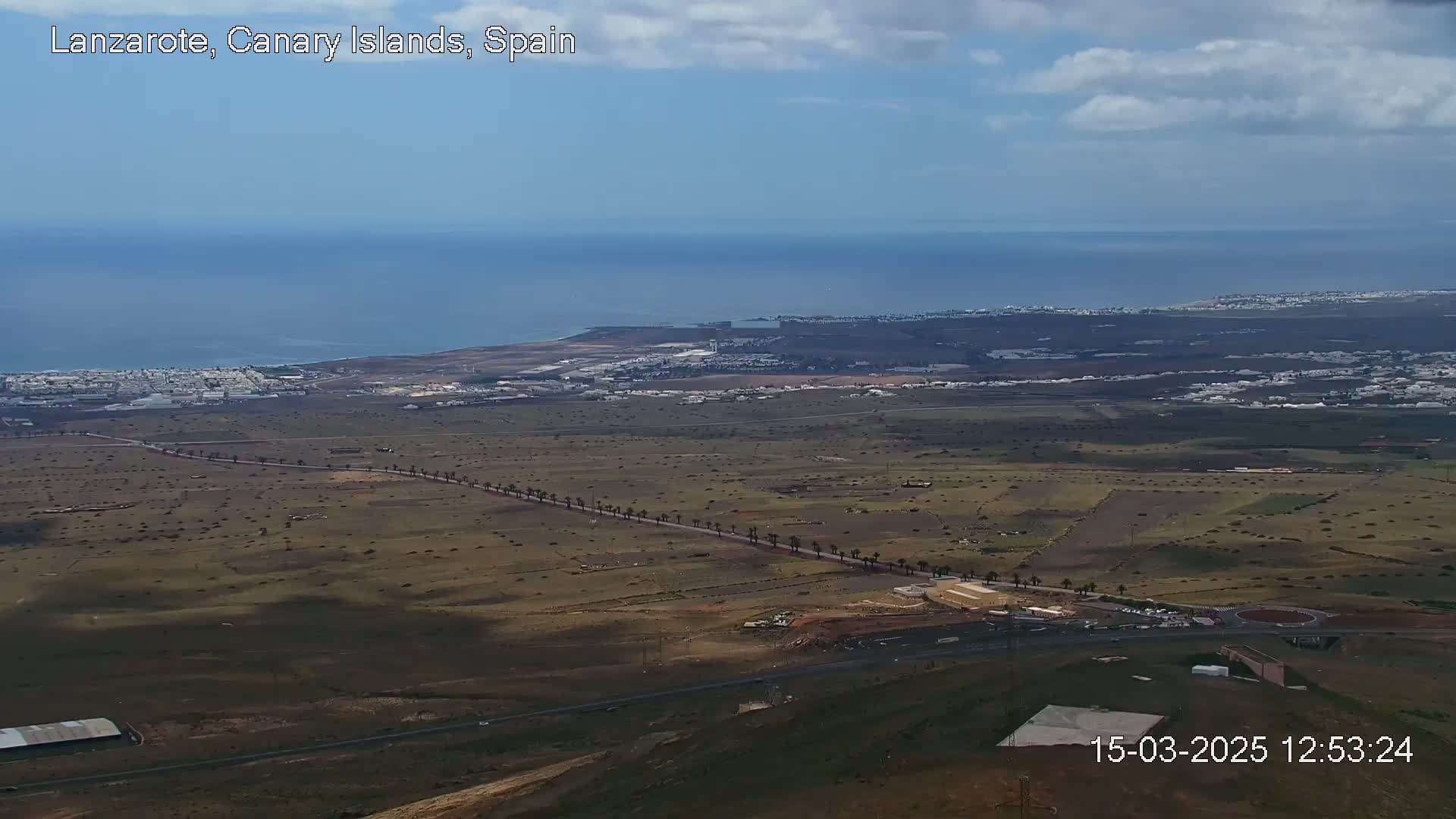 An aerial view shows a coastal area with a town, flat brown land with scattered vegetation, a long road lined with palm trees, and a partly cloudy sky.