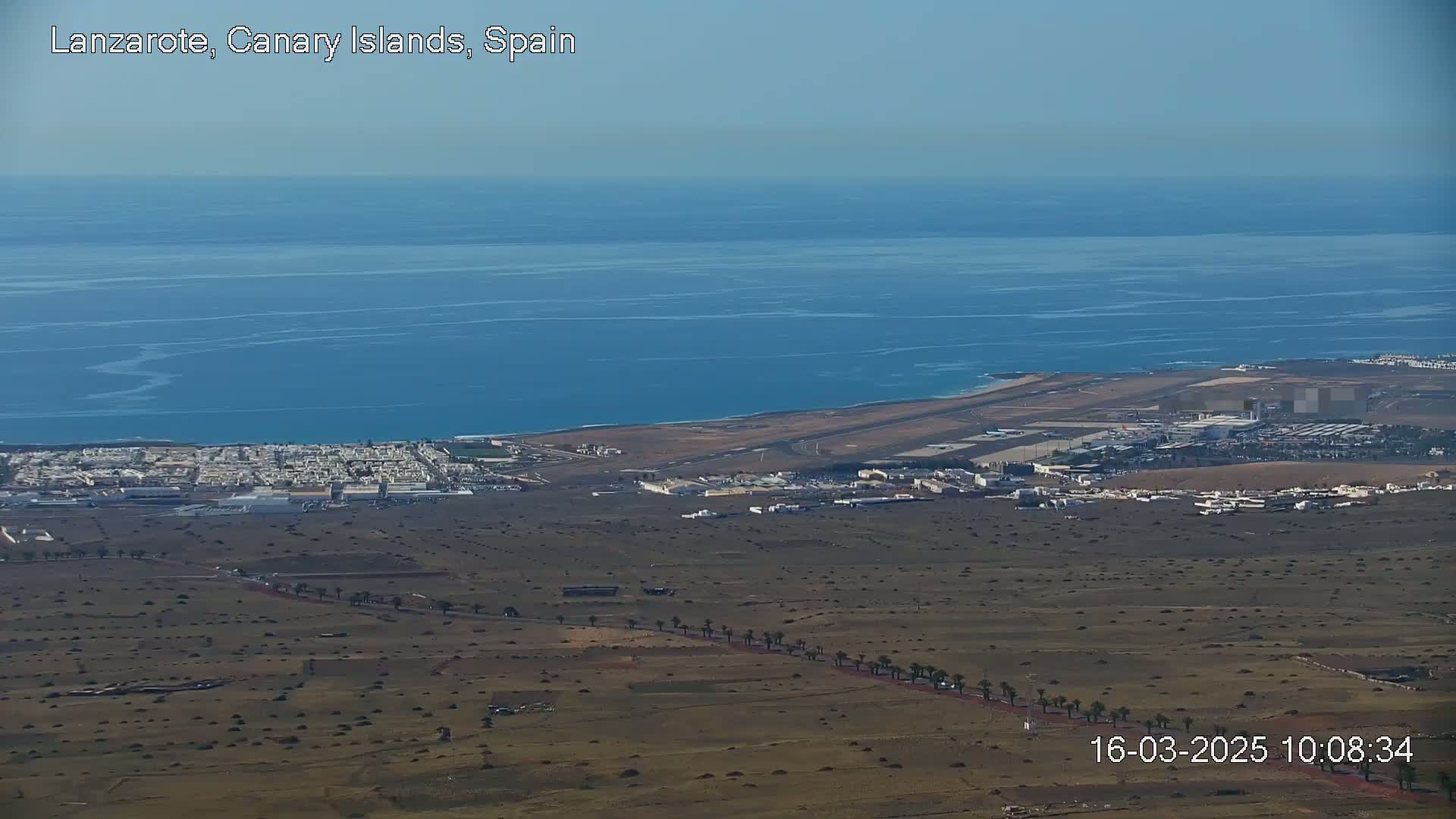 A coastal airport and town are visible below a vast expanse of calm blue ocean under a clear sky.