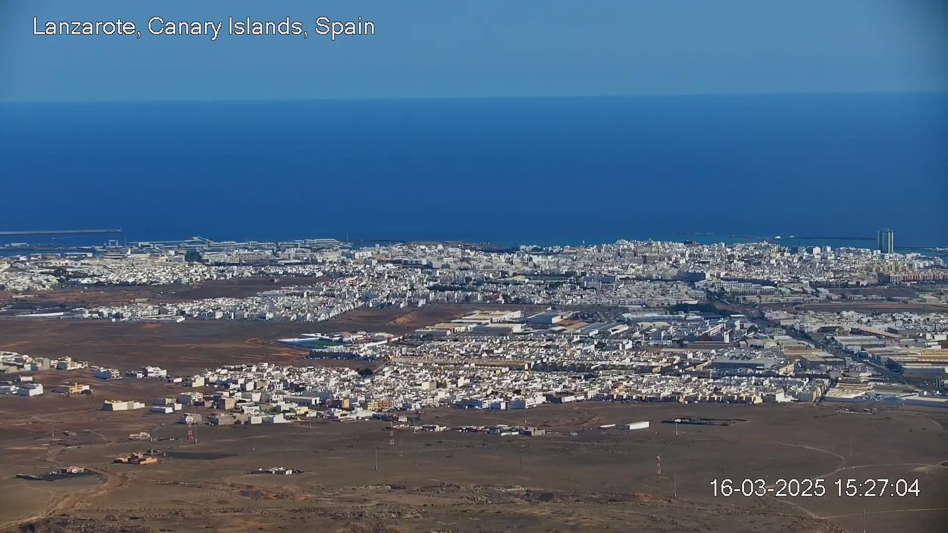 A sprawling coastal city with light-colored buildings stretches across a mostly barren landscape, under a clear blue sky and overlooking a calm, deep blue ocean.
