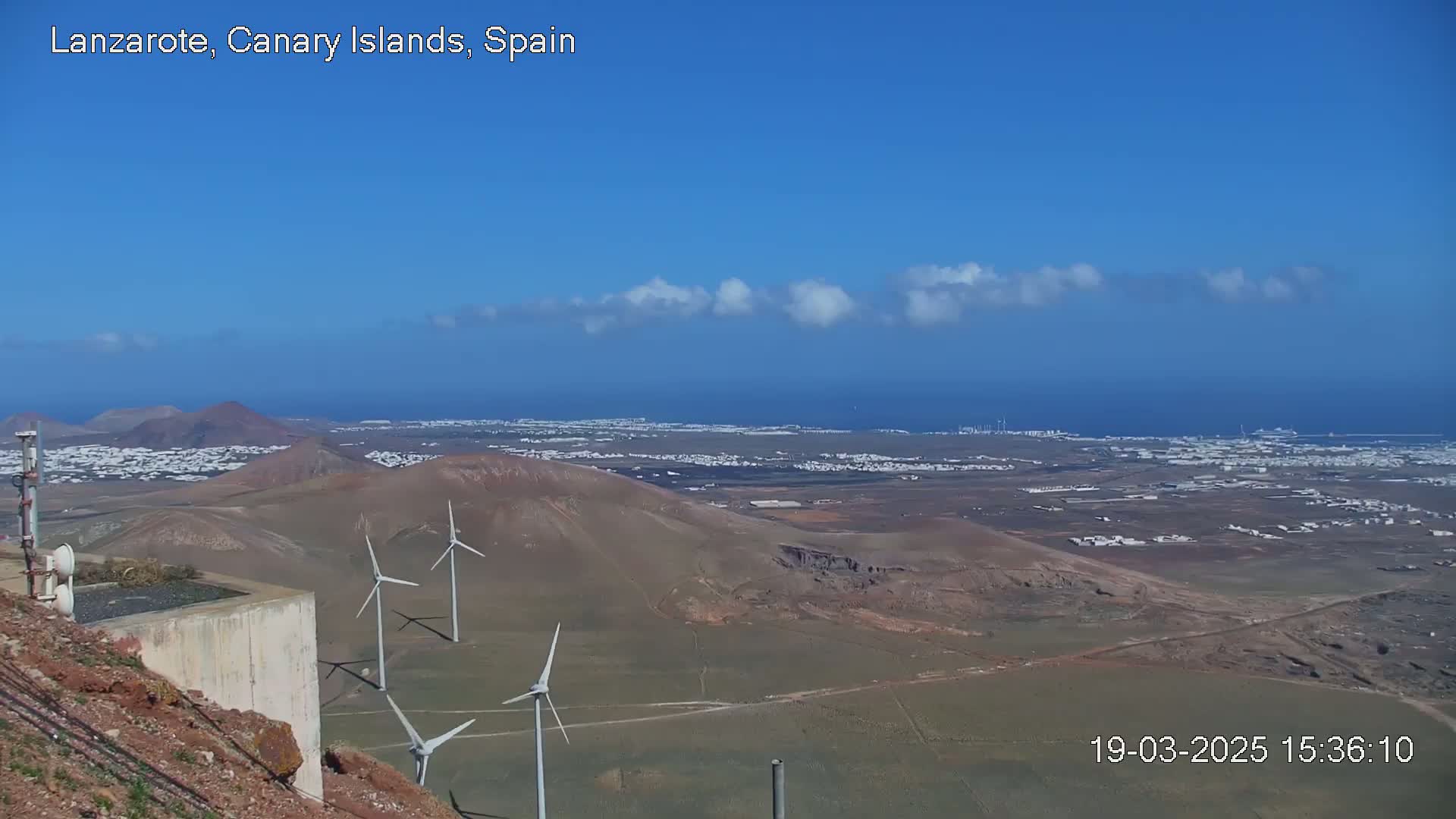 A panoramic view shows a coastal town stretching to a calm ocean under a mostly clear blue sky with a few wispy clouds, featuring several wind turbines on a hillside in the foreground.